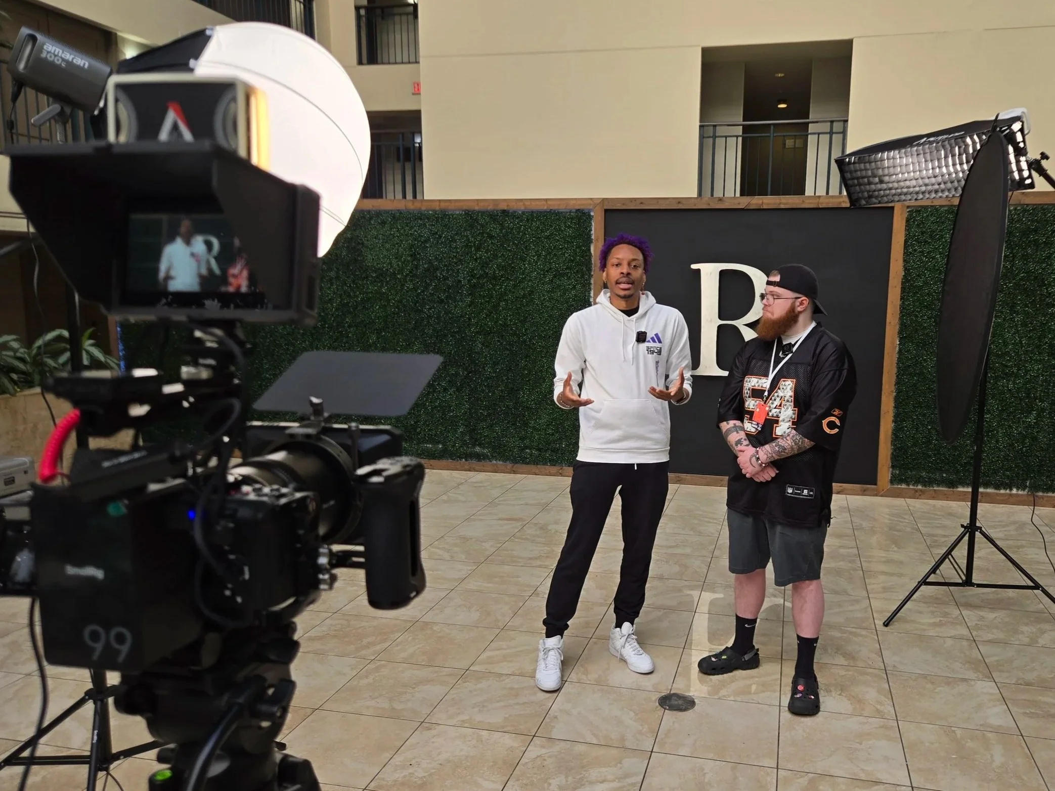 Two men standing in front of a green hedge wall with a large letter B, being recorded by professional video equipment, one speaking and the other listening.