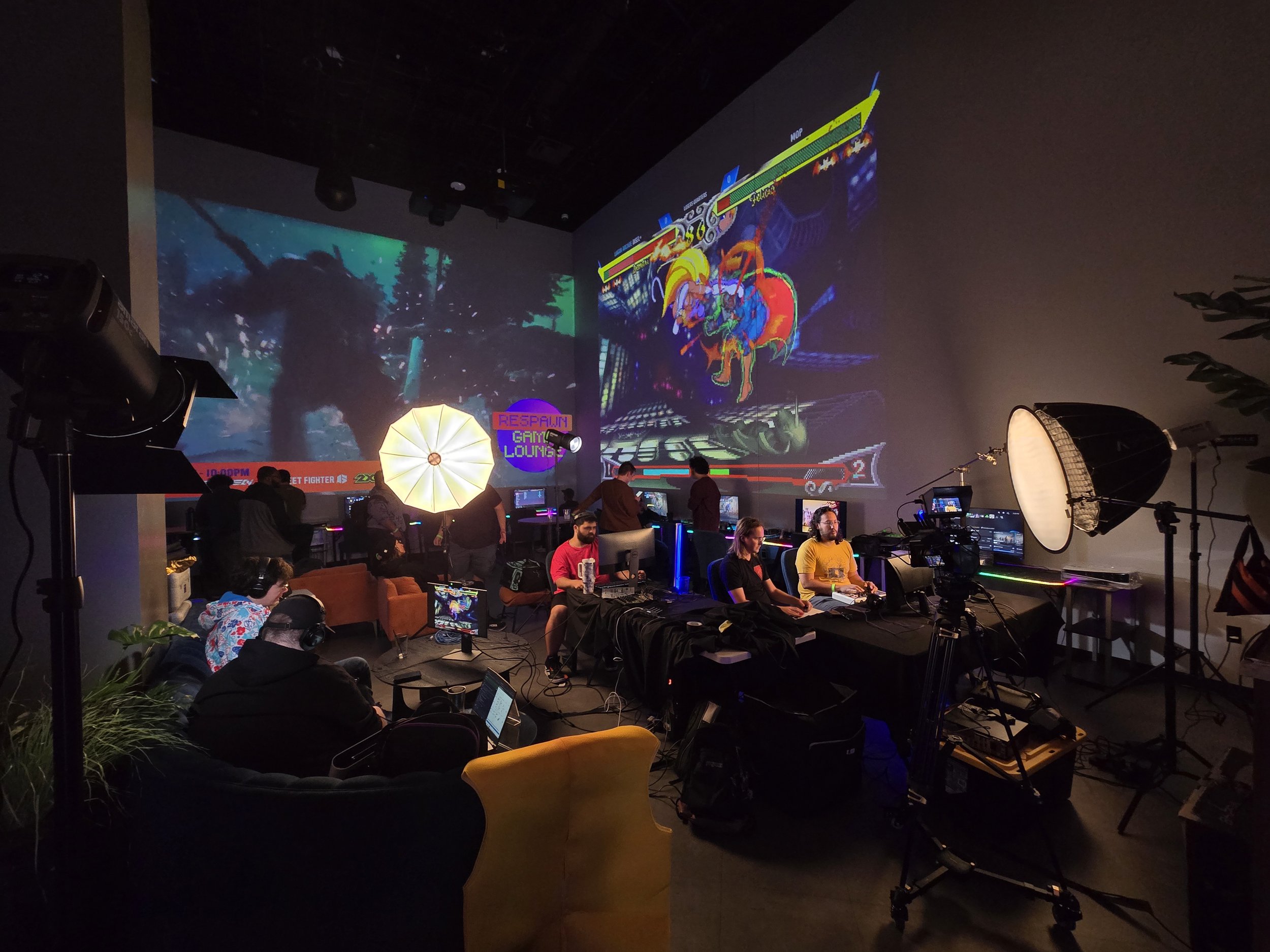 A gaming event or arcade with multiple people playing video games on various monitors. There are professional lighting and filming equipment, including softboxes and cameras. Large screens display colorful game graphics, and the setting is dark with neon lighting.