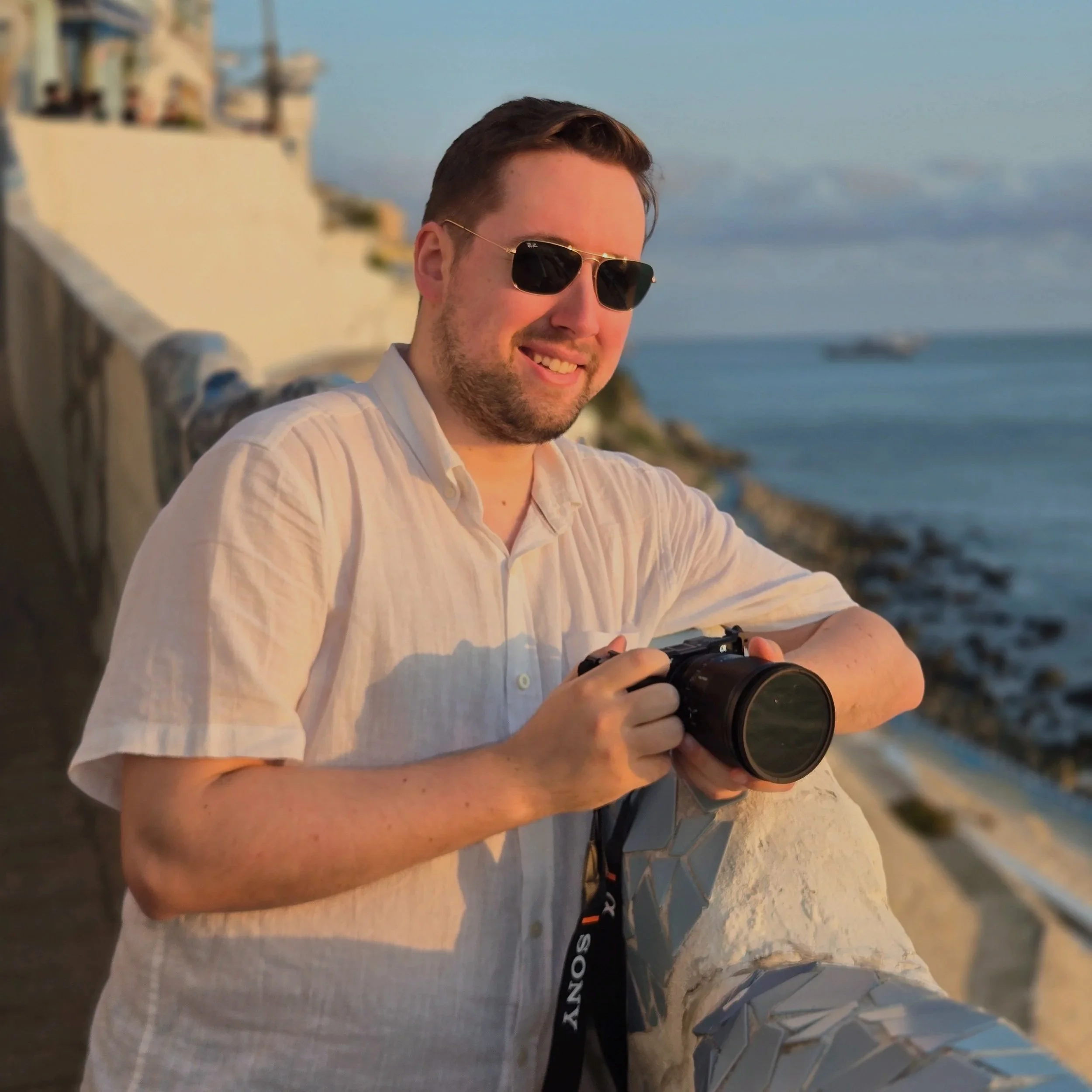 Man in sunglasses wearing a beige shirt, holding a camera, standing by a coastline during sunset.