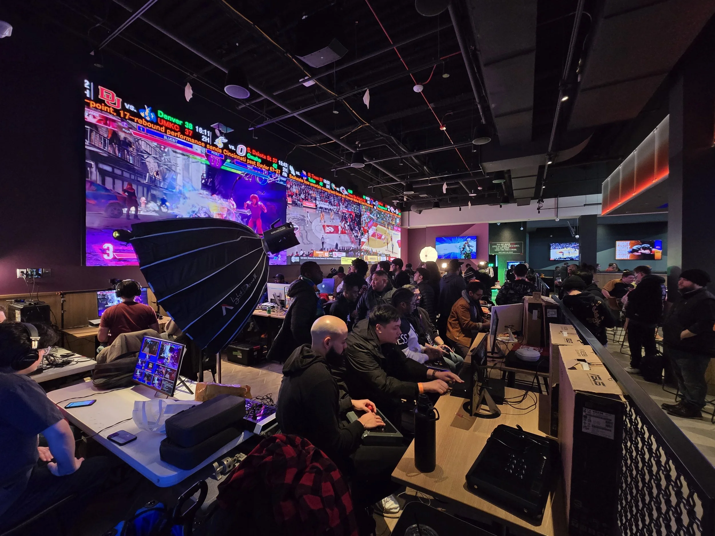 People playing video games at a gaming event or tournament, with large screens displaying gameplay, and some individuals wearing headsets and using controllers.