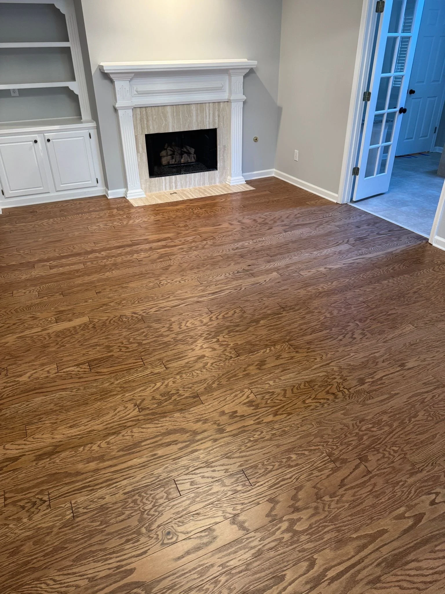 A room with hardwood flooring, a white fireplace with beige tiles, built-in white cabinets on the left, and a doorway with glass-paneled double doors on the right.