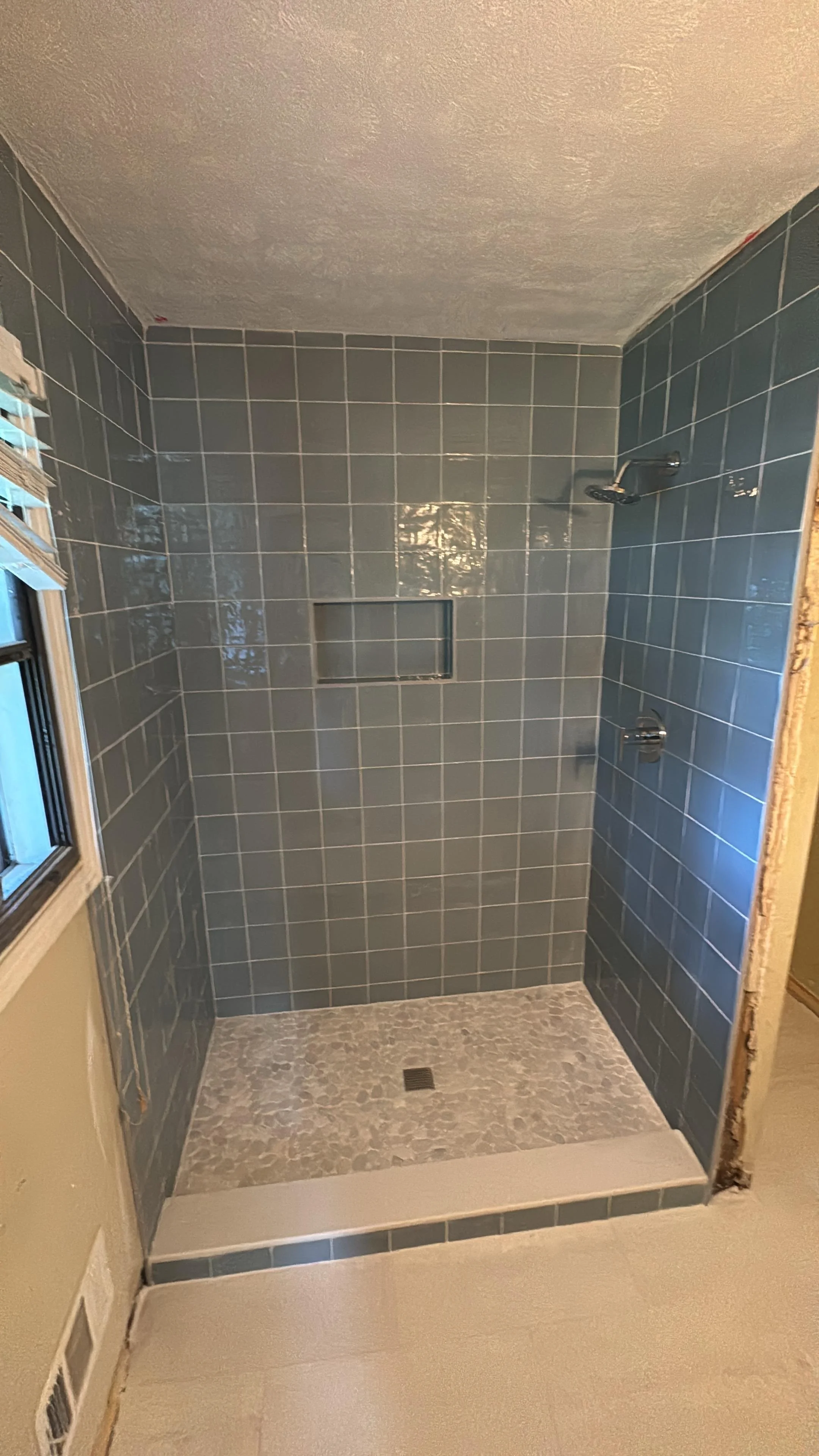 A walk-in shower with blue tiled walls, a built-in niche, a showerhead, and a control valve, with a textured ceiling and a floor drain.