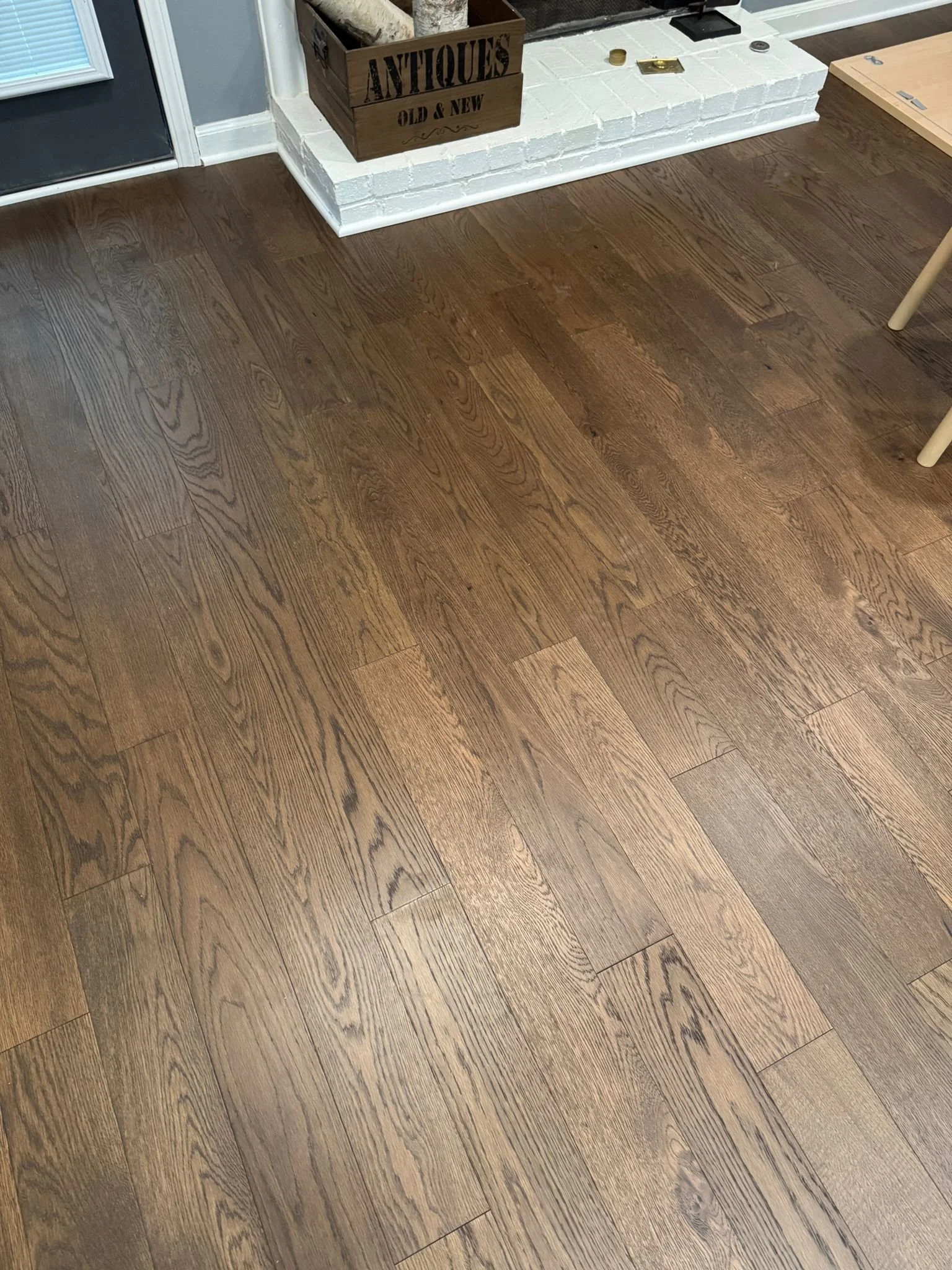 A section of hardwood floor next to a white brick fireplace with various objects on top and nearby including a wooden box labeled 'Antiques Old & New' and a piece of wood. Part of a chair leg is also visible.