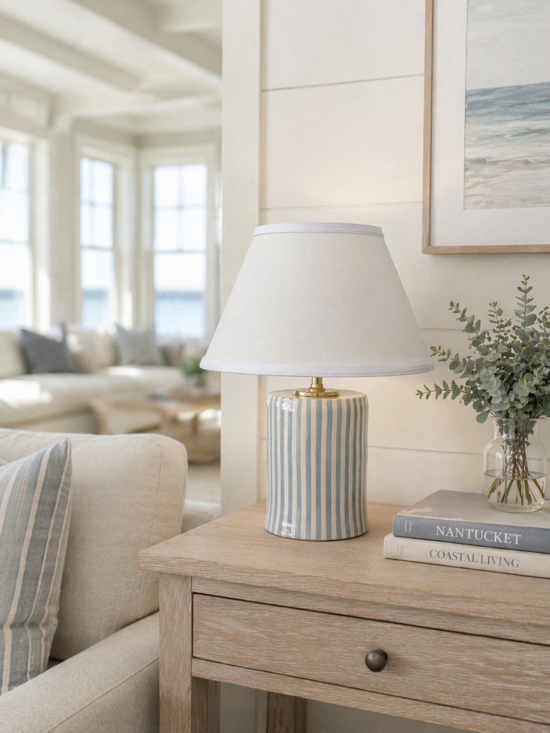 A table lamp with a striped ceramic base and white shade sits on a wooden side table next to a bronze knob, with books titled "NANTUCKET" and "COASTAL LIVING" and a small glass vase with green eucalyptus leaves in a bright, coastal living room.