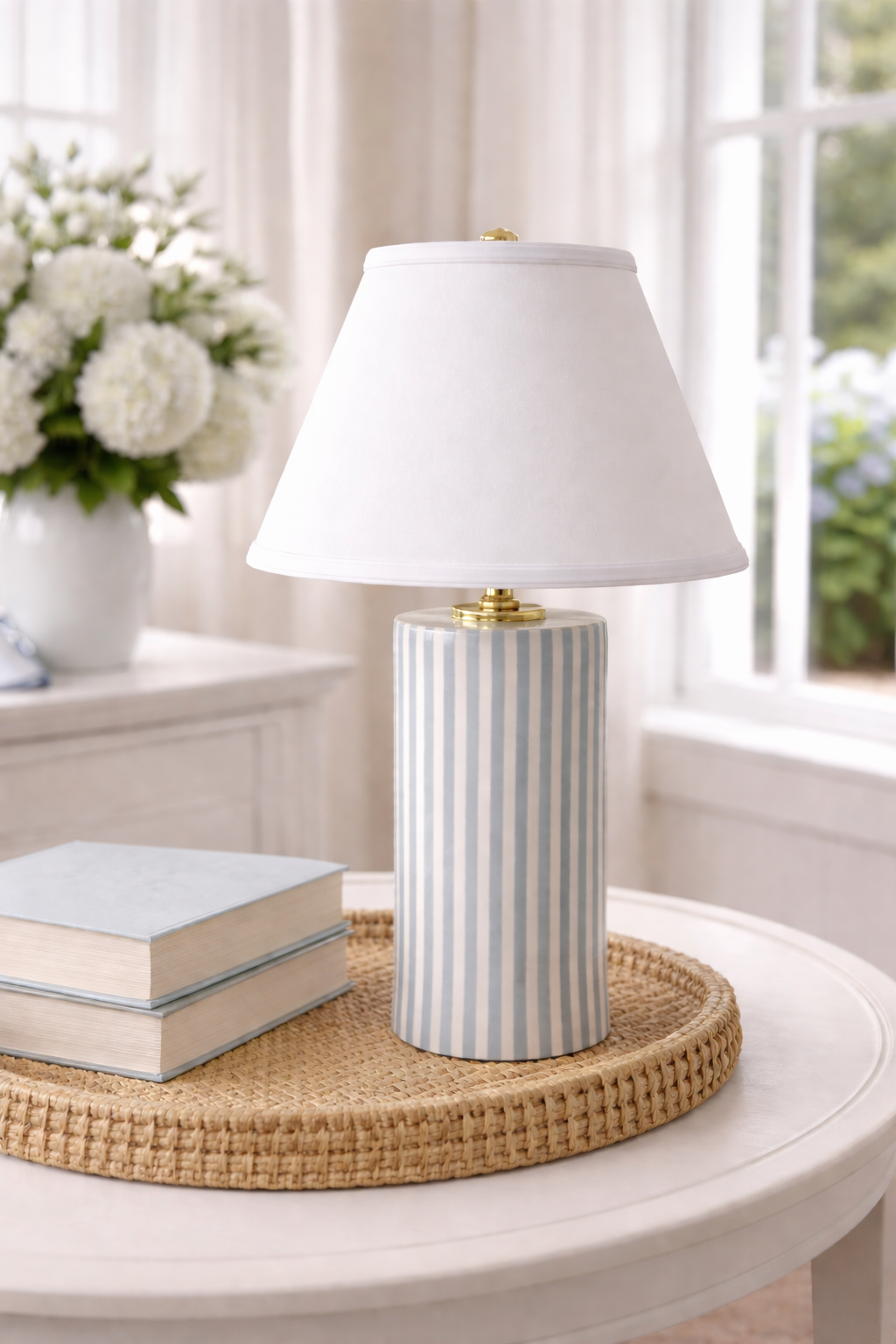 A table lamp with a white shade and a striped blue and white ceramic base, placed on a round wicker tray on a white table, with a stack of books and a vase of white flowers in the background.