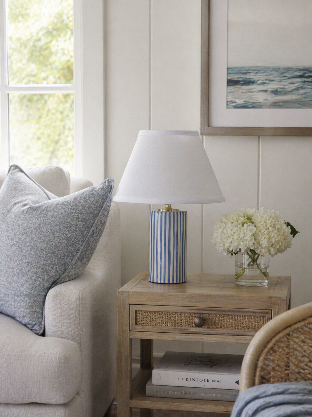 A side table with a striped ceramic table lamp, a glass vase with white hydrangea flowers, and books, next to a sofa with a patterned pillow and a window with a view of trees outside.