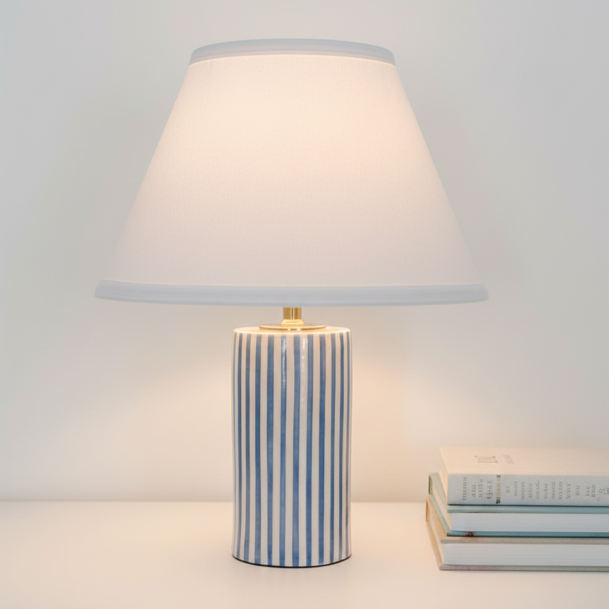 A table lamp with a cylindrical blue and white striped ceramic base and a white lampshade, located next to a small stack of books.