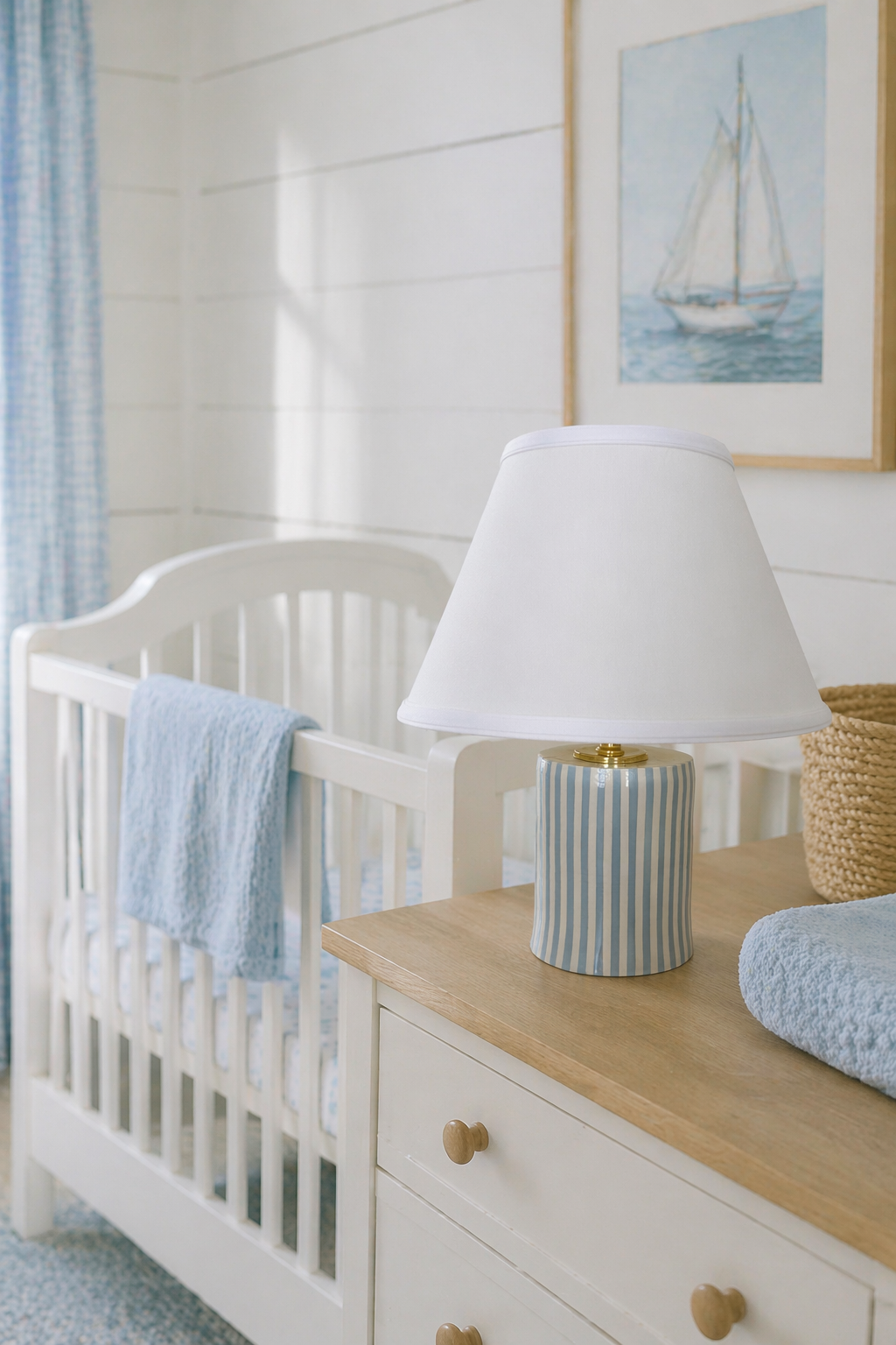 A white crib with a light blue blanket, a striped lamp, and a woven basket on a wooden dresser in a nursery with white wood-paneled walls and a framed picture of a sailboat.