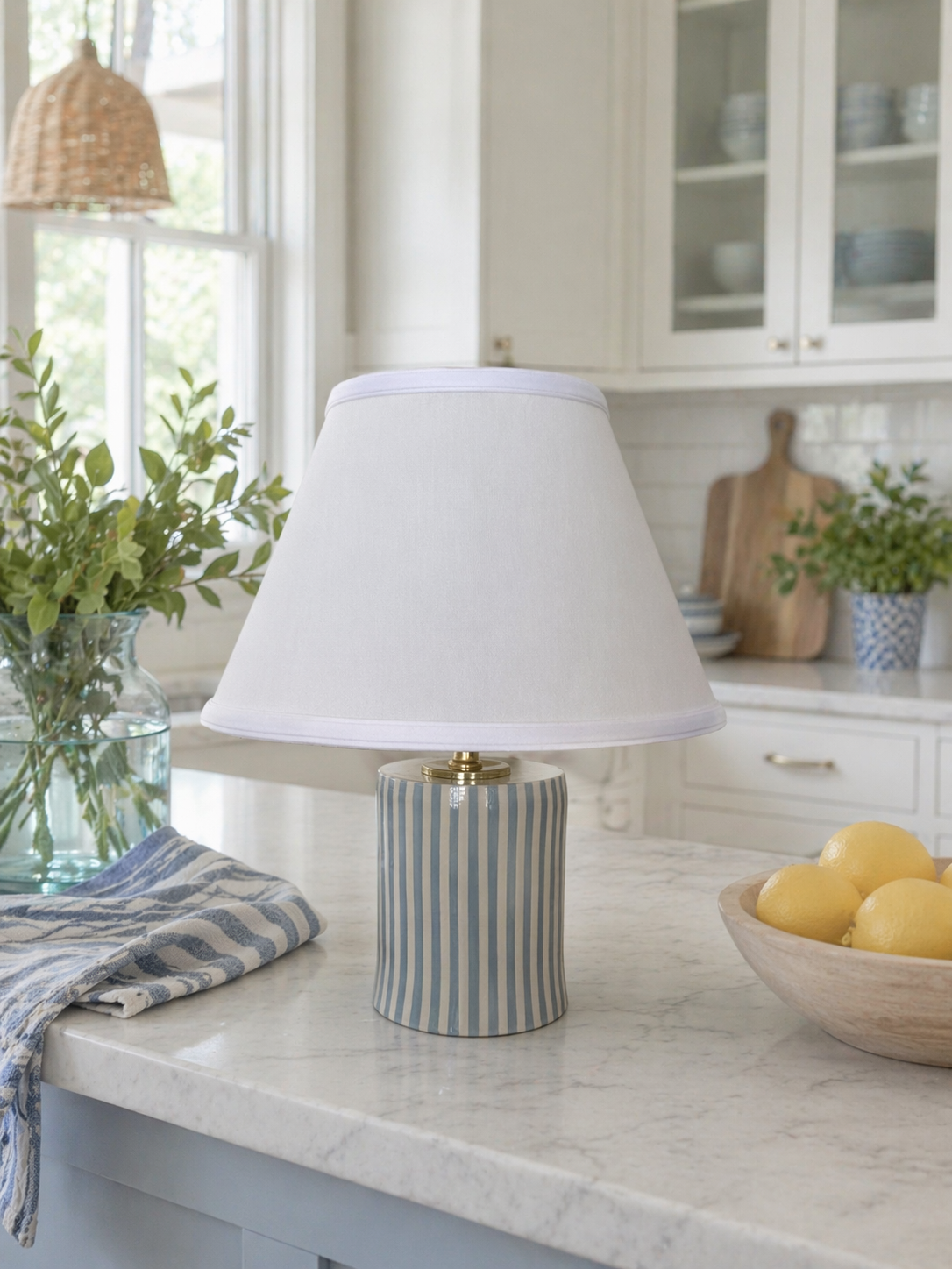 A ceramic table lamp with a white lampshade and striped blue and white base on a kitchen marble countertop, with a bowl of lemons and green plants nearby.