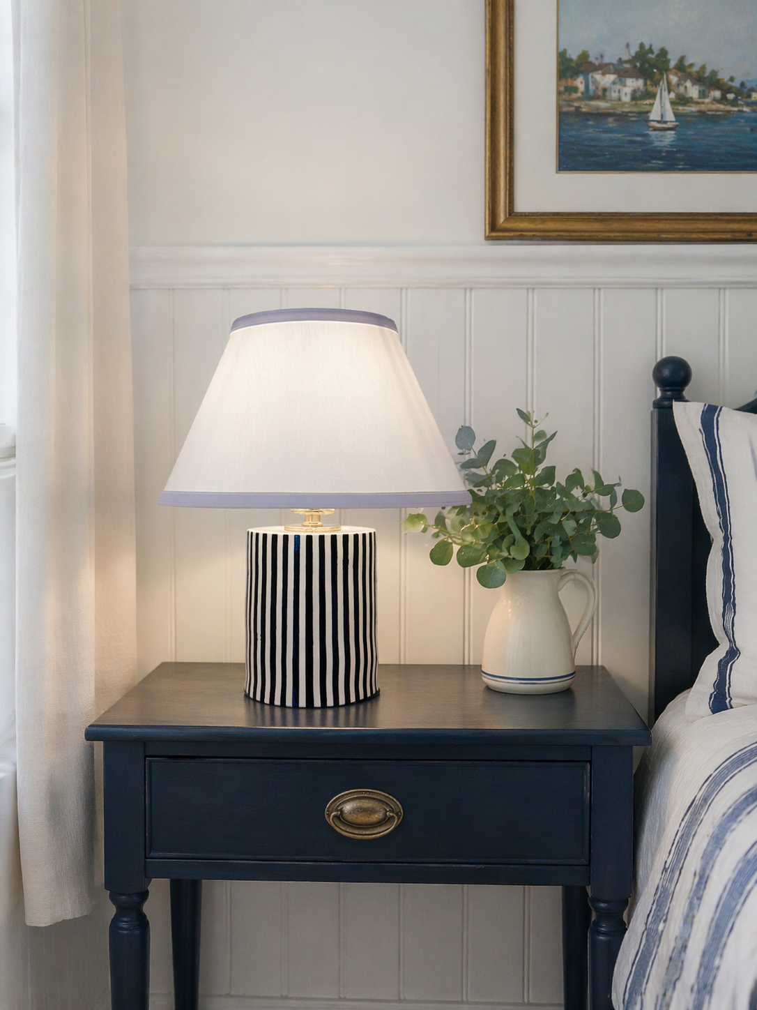 A bedside table with a black and white striped lamp, a white pitcher filled with green foliage, a framed painting of a coastal scene, and a partial view of a bed with a striped pillow.