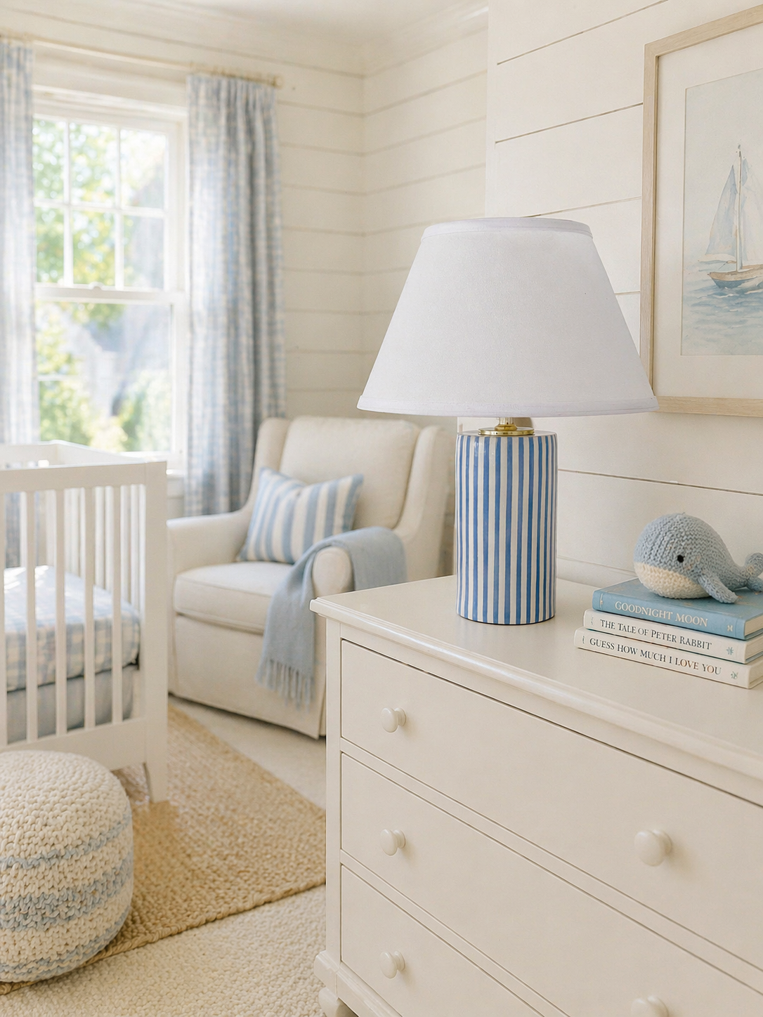 Cozy nursery with white wooden walls, a white crib, a beige armchair with a blue and white striped pillow, a white dresser with a blue striped lamp, a crocheted whale toy, and stacked children's books, with a window letting in natural light.