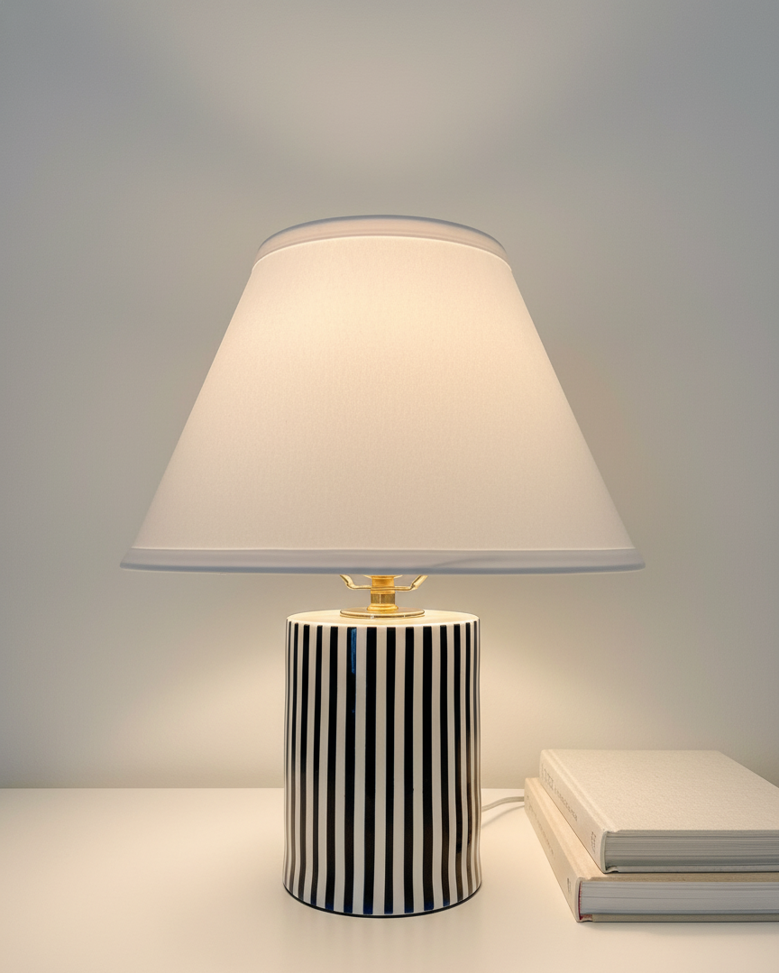 A table lamp with a black and white striped ceramic base and a white lampshade, positioned on a white table next to two stacked books.
