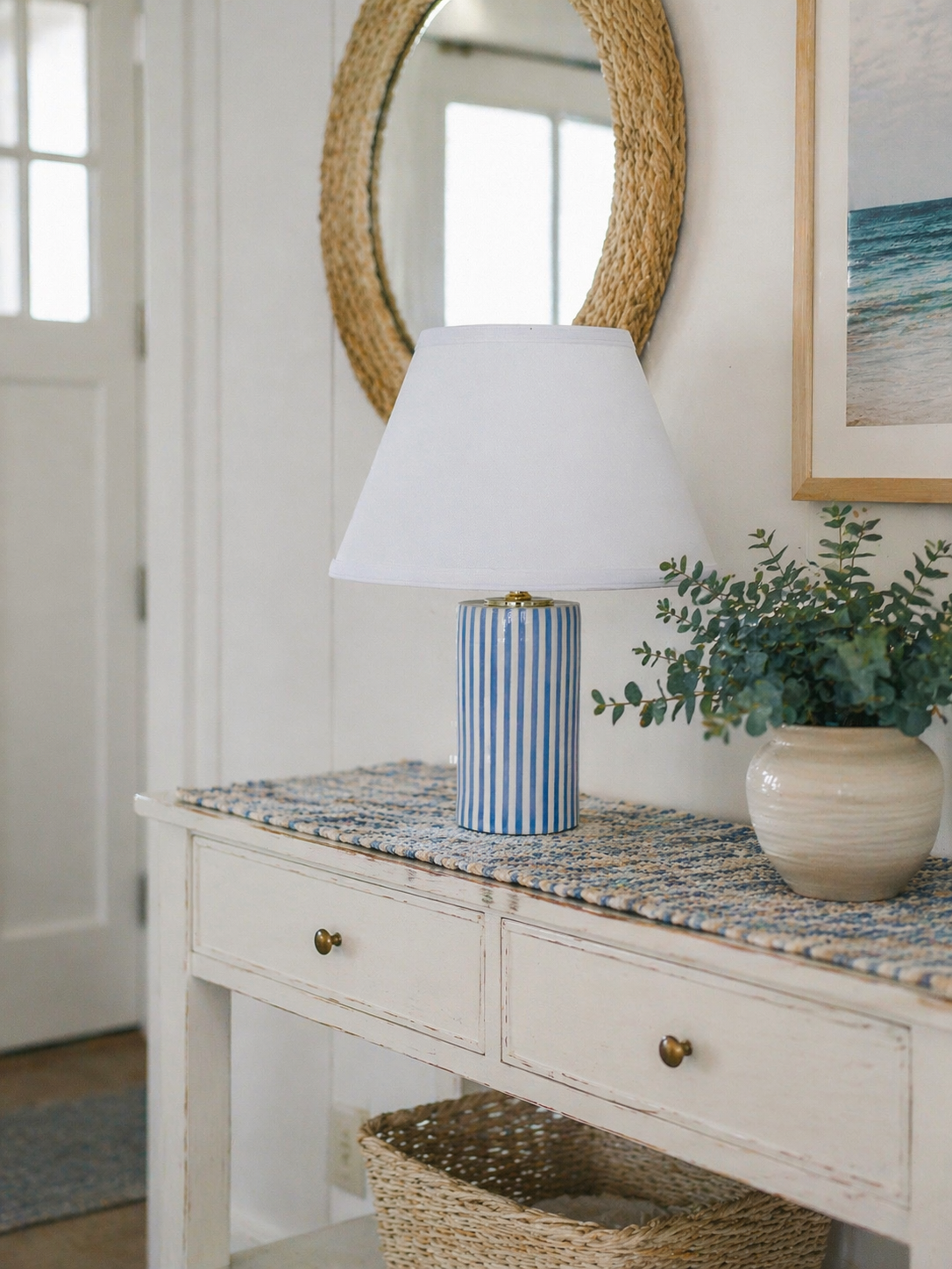 A white wooden console table with two drawers, topped with a blue and white striped table lamp and a white ceramic pot with greenery. A round, woven mirror hangs above and to the left of a beach-themed framed picture on the wall.
