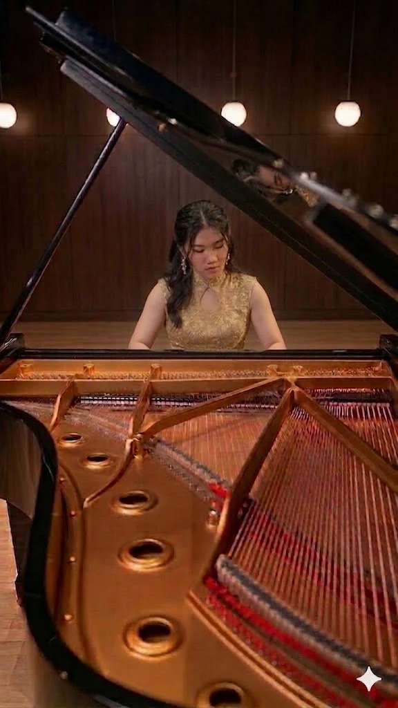 A woman in a gold dress playing a grand piano in a music hall with wooden walls and hanging round lights.
