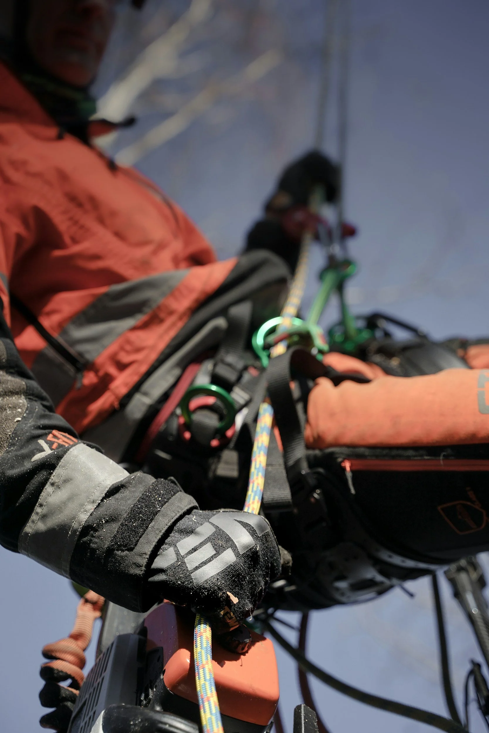 A person in outdoor gear adjusting climbing or rappelling equipment, with a snowy background and a clear sky.