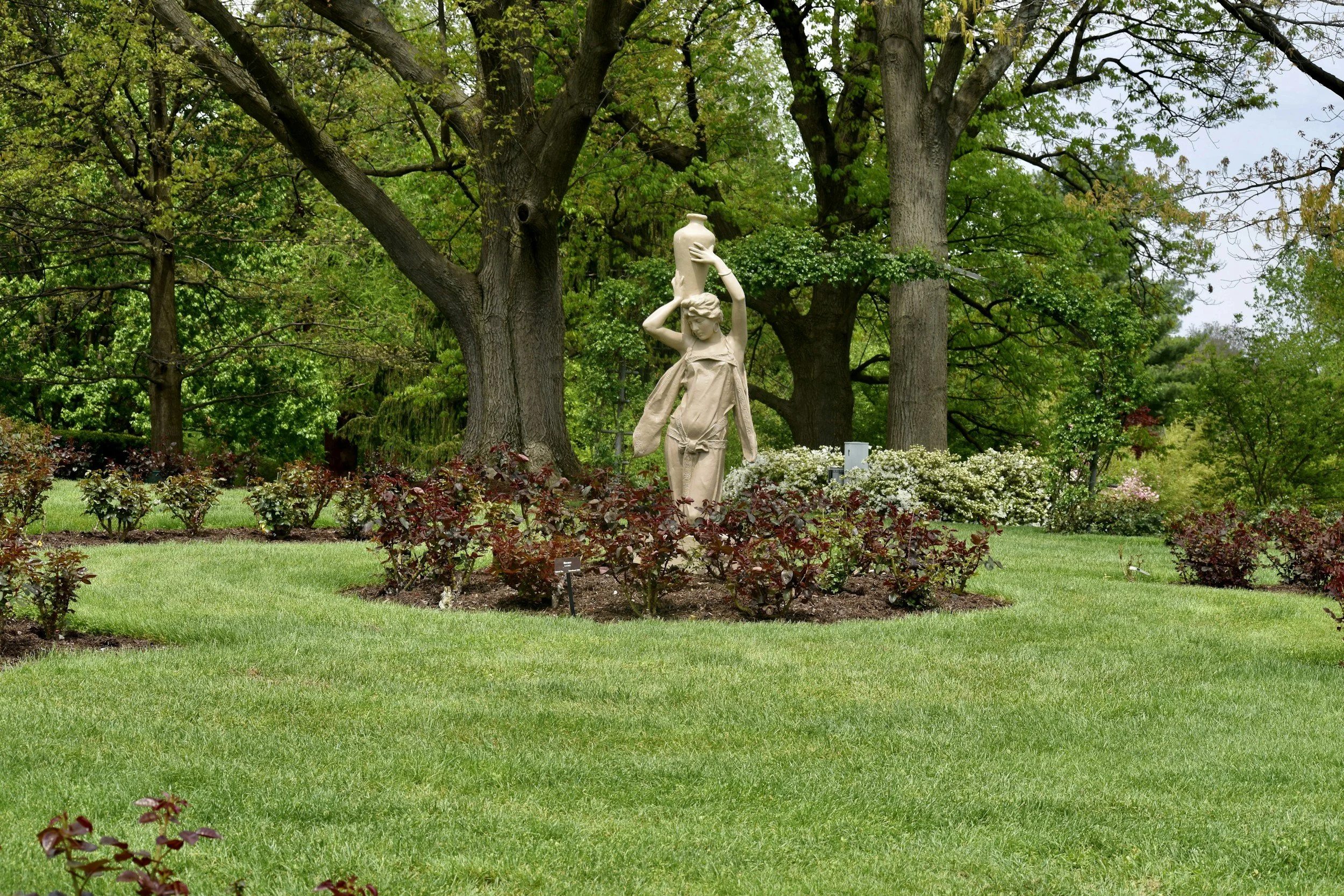 Statue of a woman holding a vase, set in a lush garden with large trees, blooming bushes, and well-manicured grass.