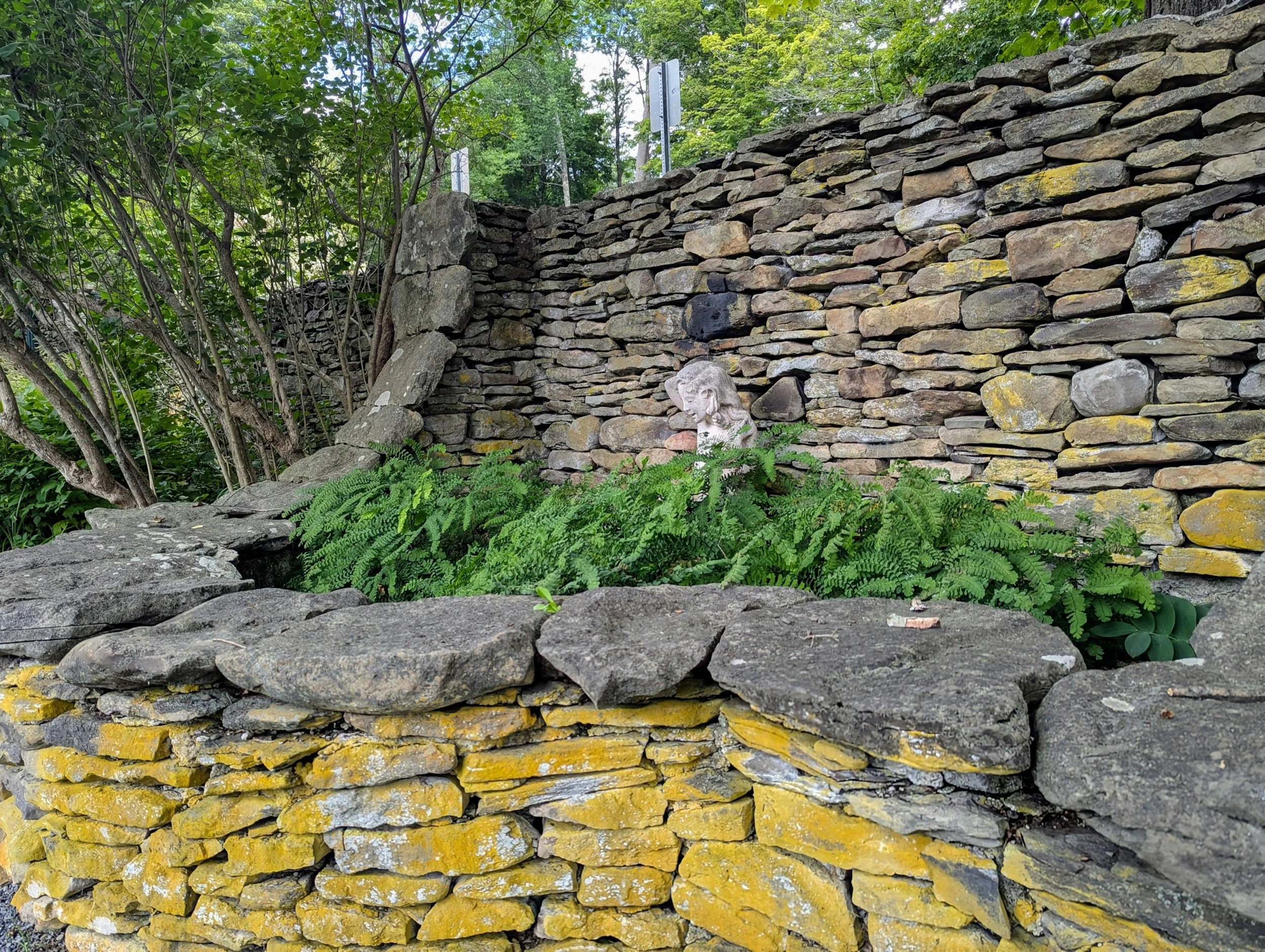 Stone wall with greenery and a small ceramic or stone bust of a child's head among plants.