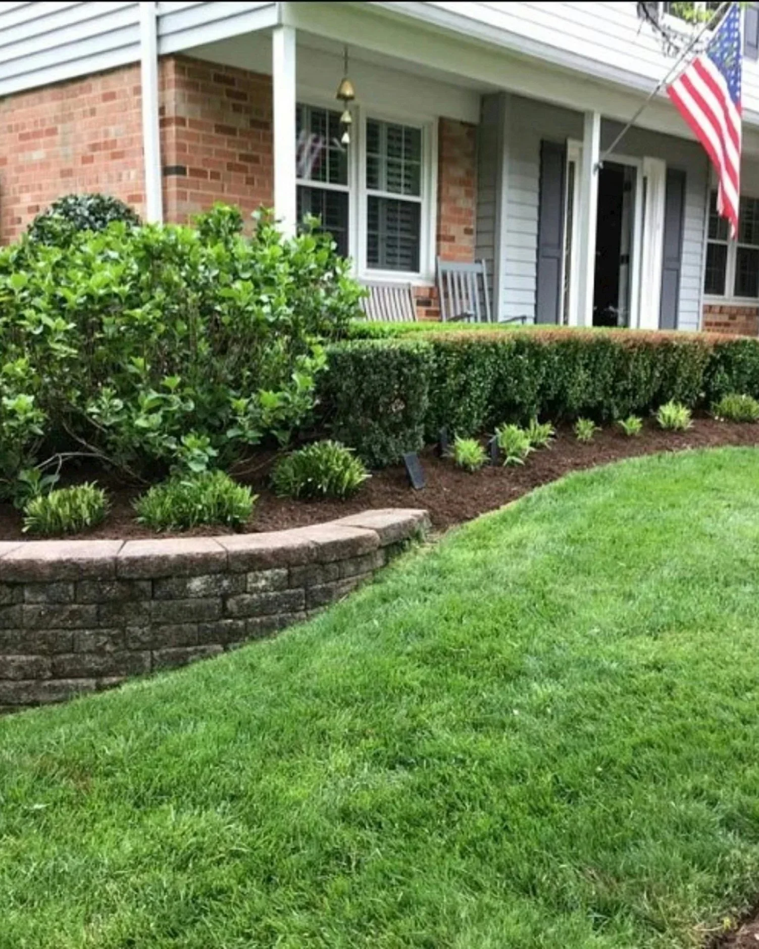 A well-maintained front yard with a lush green lawn, neatly trimmed bushes, and flowering plants, in front of a brick and siding house with American flag hanging on the porch.