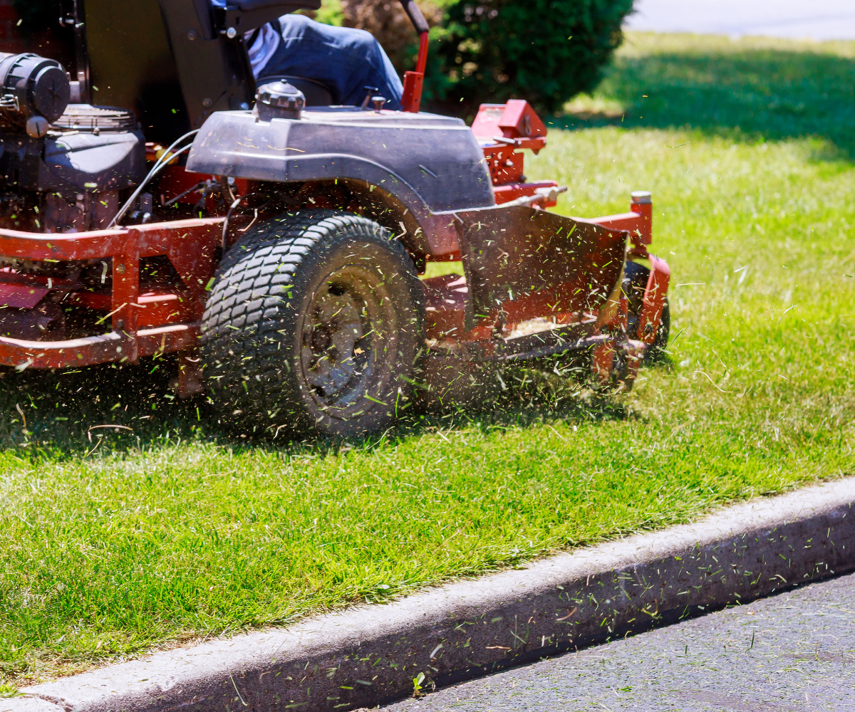 A person riding a red lawn mower cutting grass along a curb during daytime.