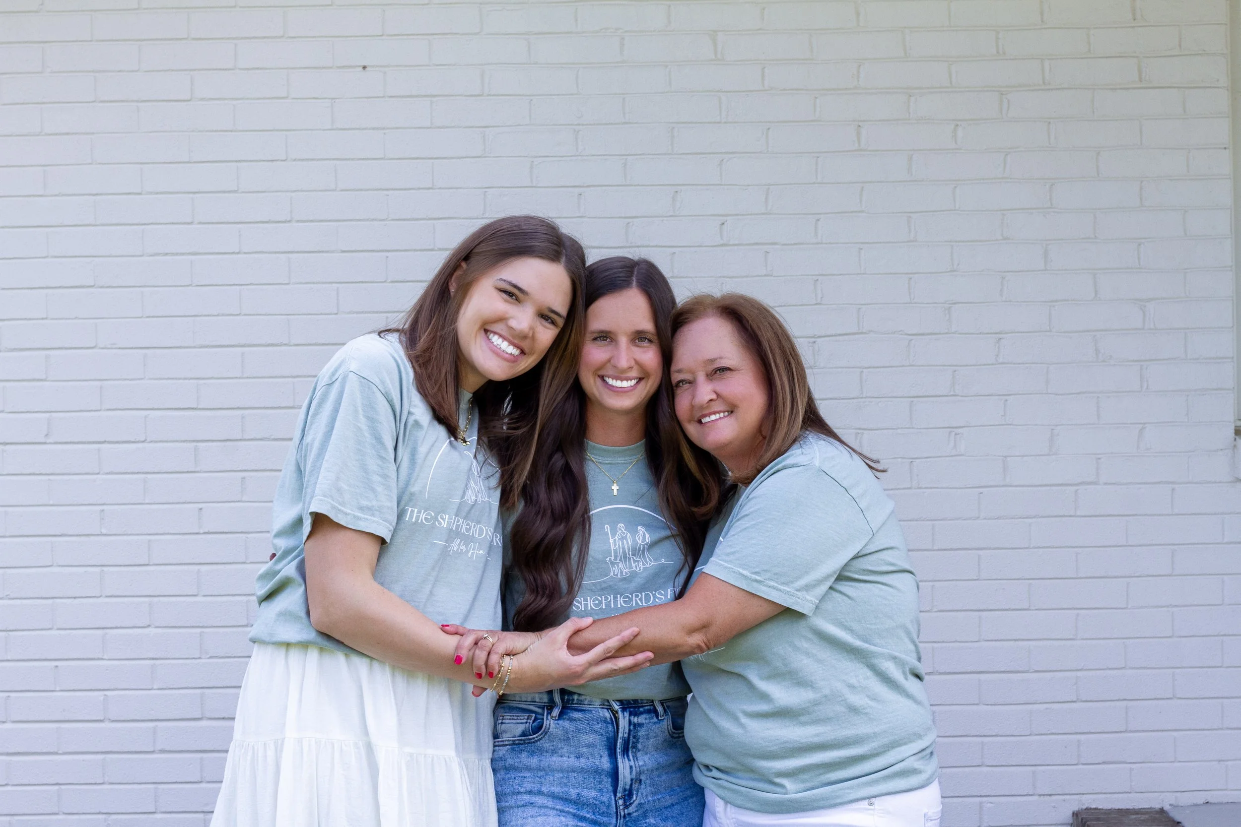 Three women hugging and smiling in front of a white brick wall.