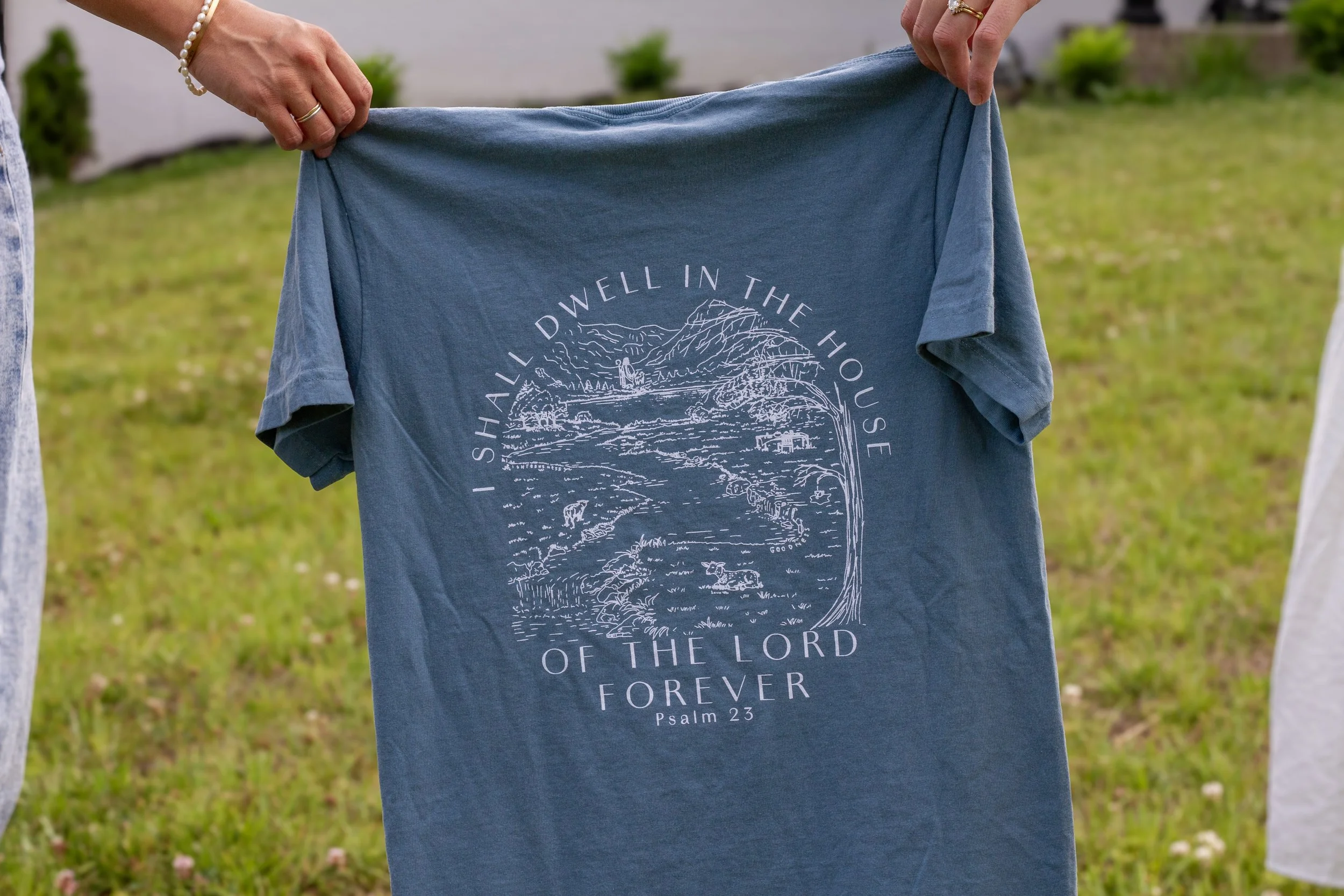 Two people are holding a blue T-shirt with a white graphic and text that says, 'Shall I dwell in the house of the Lord forever? Psalm 23.' Their hands are visible, and they are standing outside on grass with some plants in the background.