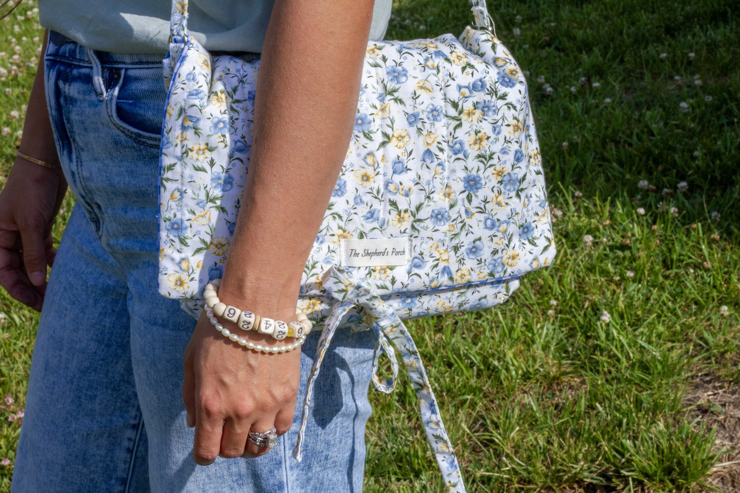 Close-up of a person wearing a floral-patterned backpack, a beaded bracelet, and rings, standing on grass in a sunny outdoor setting.