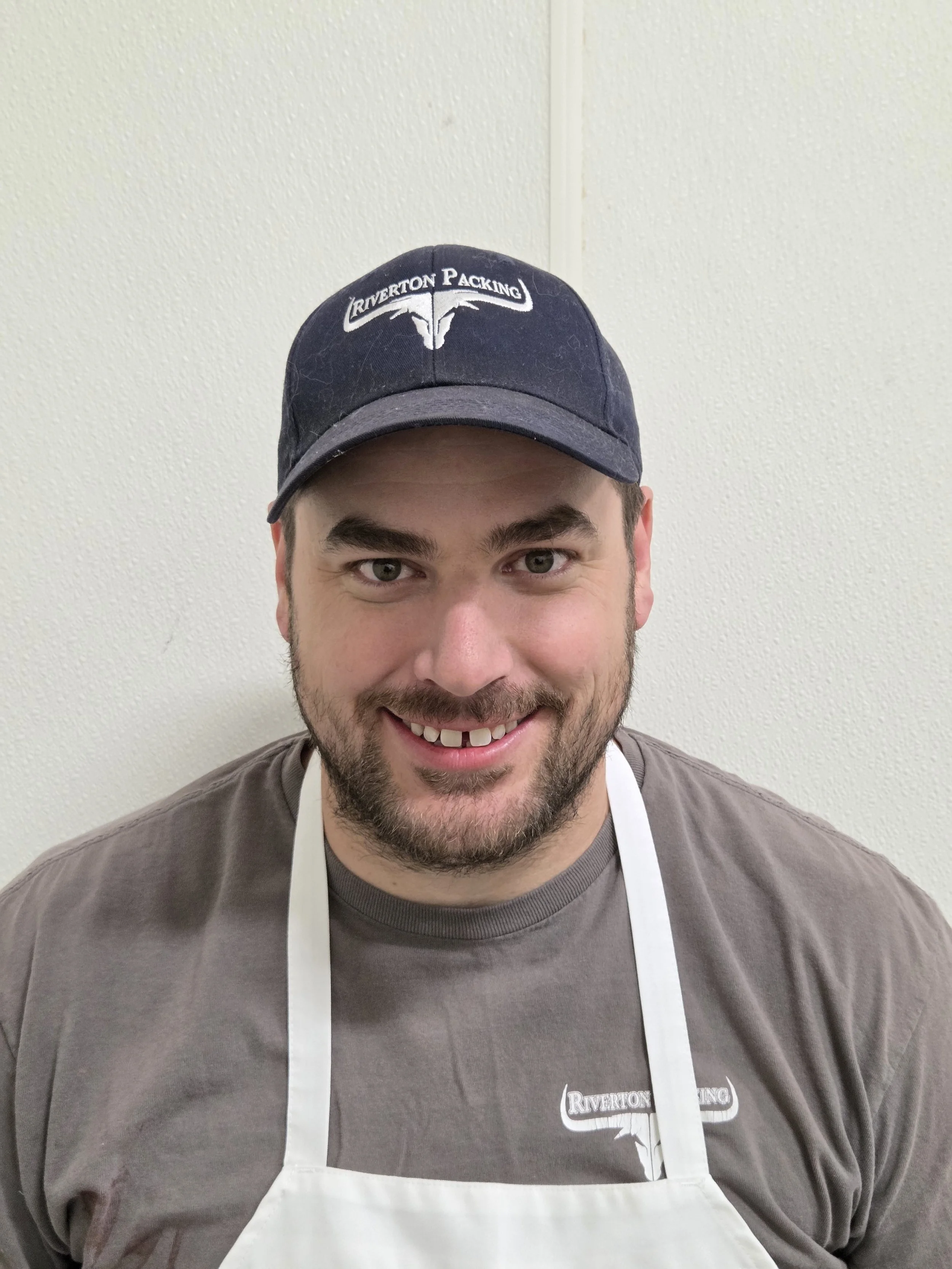 A man with a beard smiling, wearing a black baseball cap with "Riverton Packing" logo and a gray shirt, standing against a plain white wall.