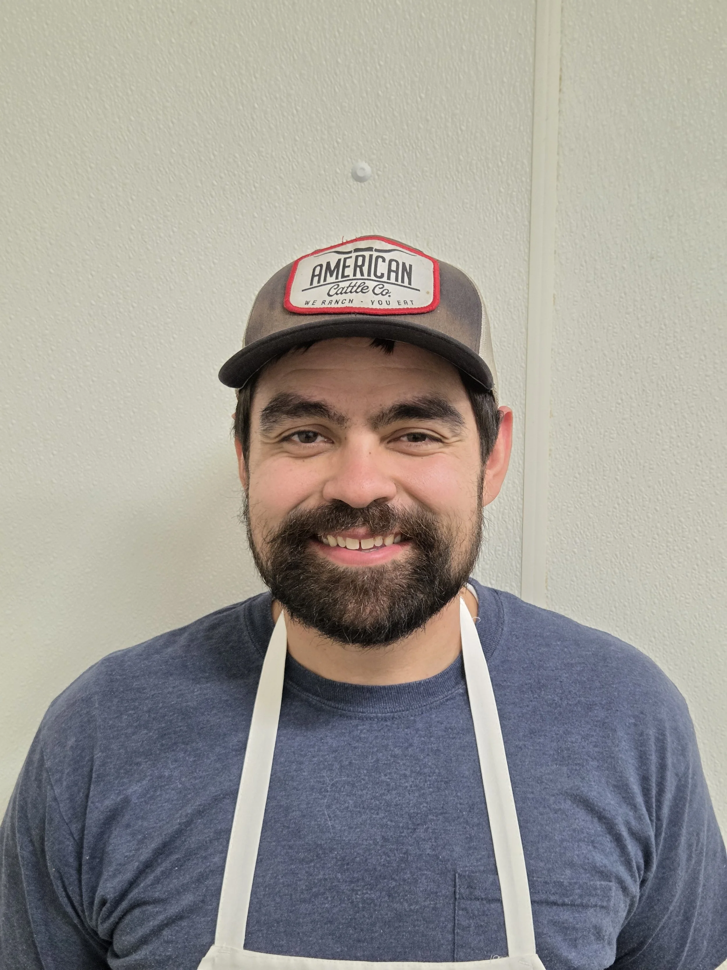 A man with a beard smiling, wearing a cap with a red and white logo reading 'American Cattle Co.' and a navy blue shirt, standing against a beige textured wall.
