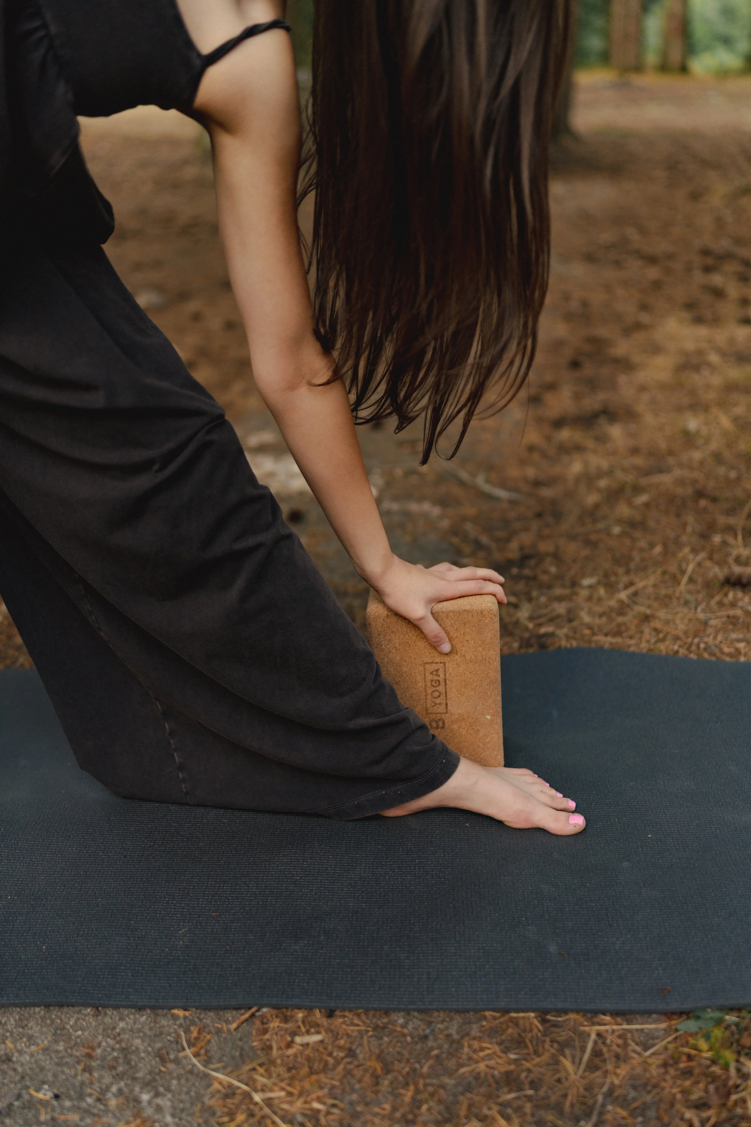 Shaina practicing triangle pose on a black yoga mat, block under her hand for support, outside on grass.