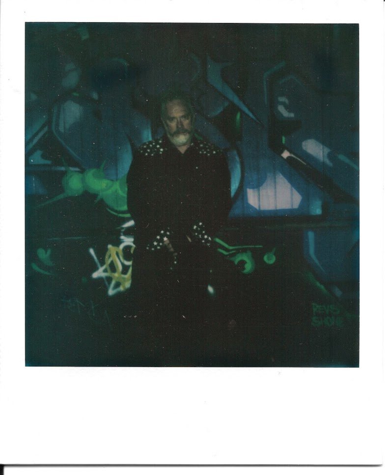 dark, far off shot of a salt-and-pepper bearded man in a black western shirt leaning against a dark graffiti-covered wall