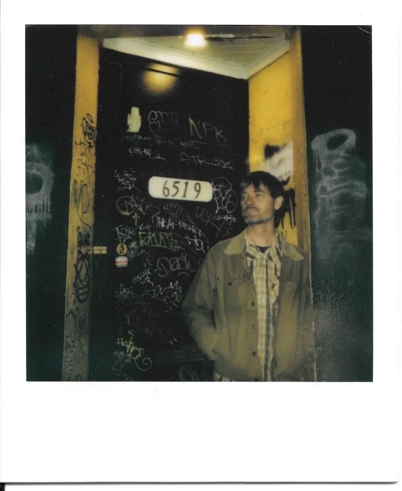 A short haired guy in a brown trucker's jacket over a plaid snap shirt with a close-trimmed beard leaning with his hands in his jacket pockets in the doorway of a graffiti-covered building with the house number 6519 on the black door