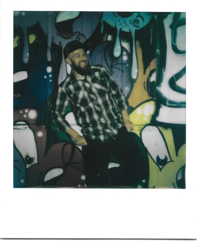 a guy with a baseball hat, long beard, and plaid flannel with the sleeves pulled up making a cheesy, toothy smile at the camera with his hands on his hips and one leg raised in front of a graffiti-covered wall