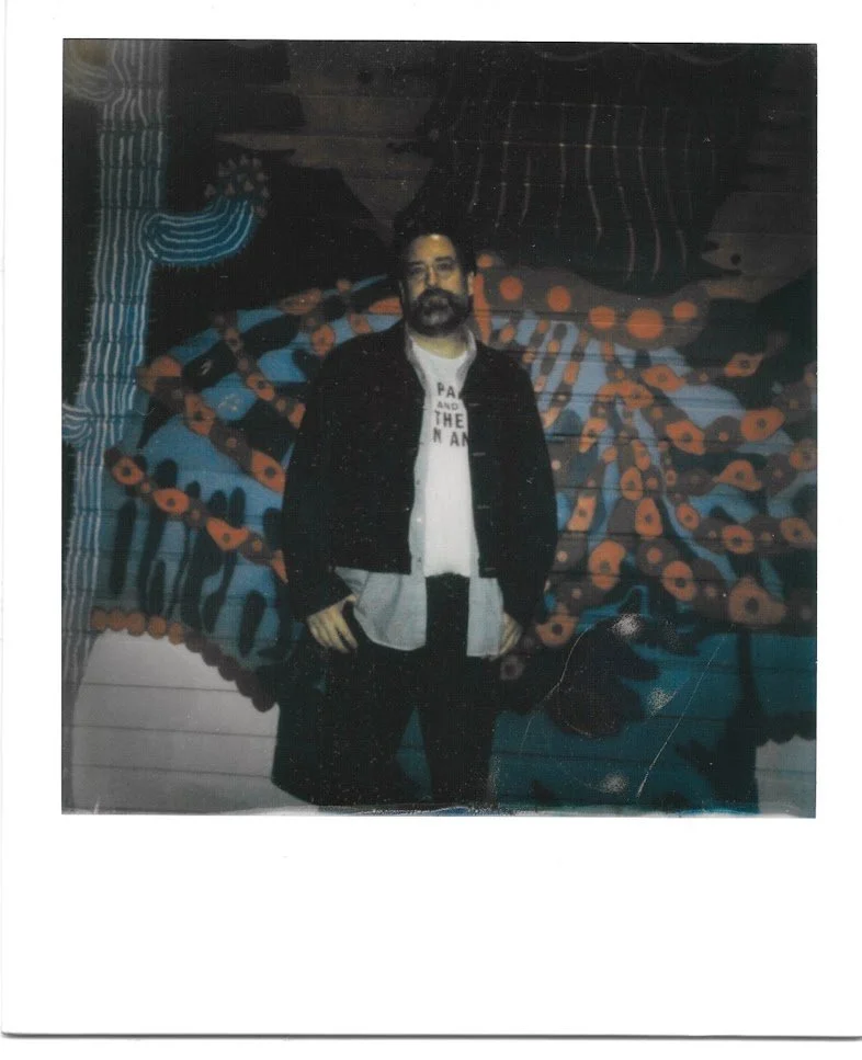 far away shot of a bearded man in a sherpa jacket over a white t-shirt with illegible black lettering standing in front of a graffiti-covered wall with his hands at his side