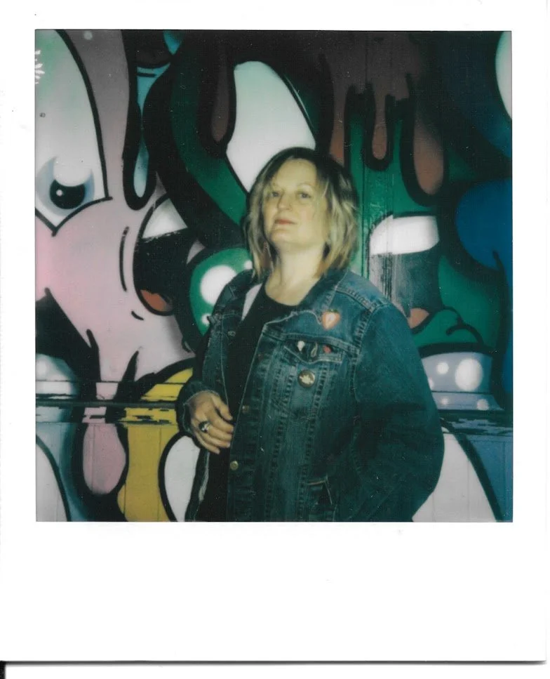 Polaroid photo of a young beautiful blonde woman in a blue denim jacket leaning against a graffiti-covered wall looking awesome