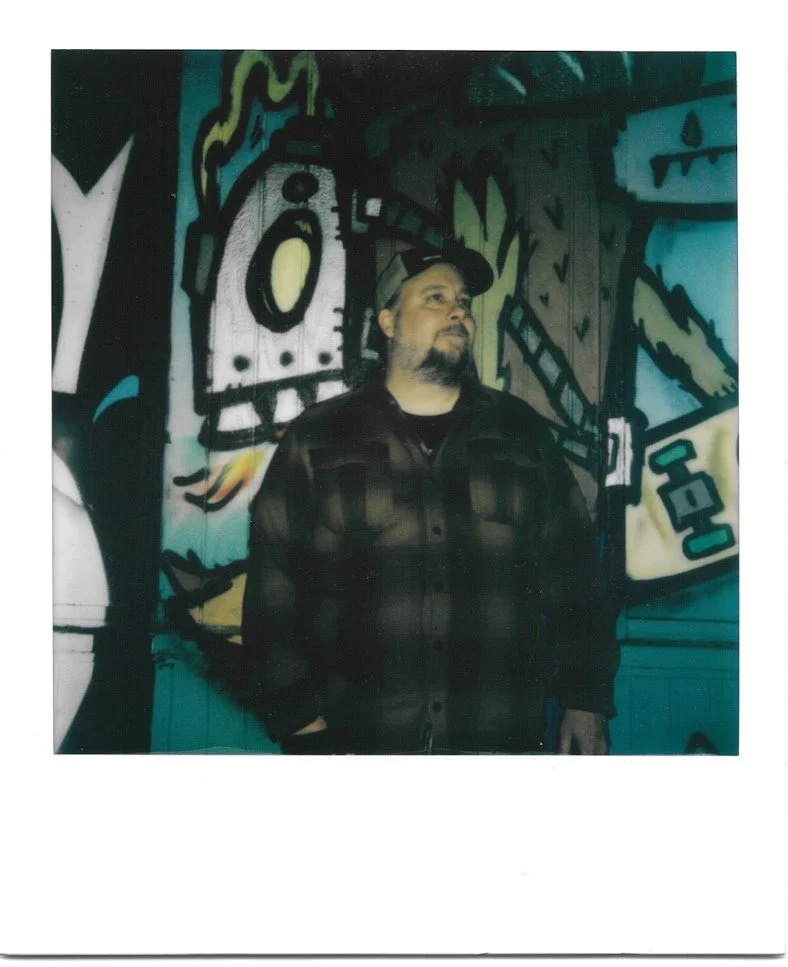 Polaroid photo of a middle aged man in a plaid flannel shirt and trucker cap with a mustache and goatee standing in front of a graffiti-covered wall at night looking to his left above the camera