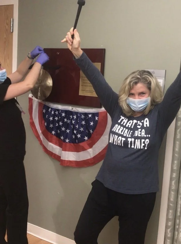 Woman celebrating in front of a plaque and American flag bunting, wearing a face mask and a shirt that says 'That's a horrible idea... What time?' while another person is removing a plaque from the wall.