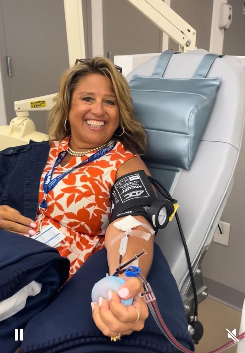 Woman donating blood in a medical setting, smiling and lying on a bed with a blue pillow, wearing a floral orange and white top and jewelry.