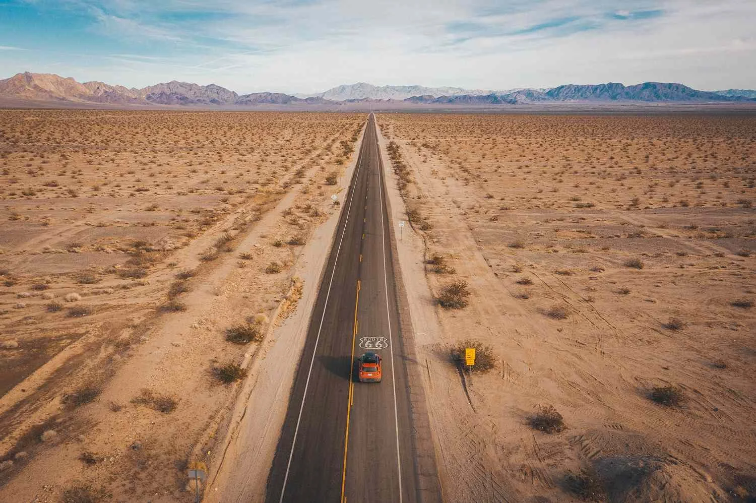 An aerial view of a straight road through a desert landscape with sparse vegetation and distant mountains under a partly cloudy sky.