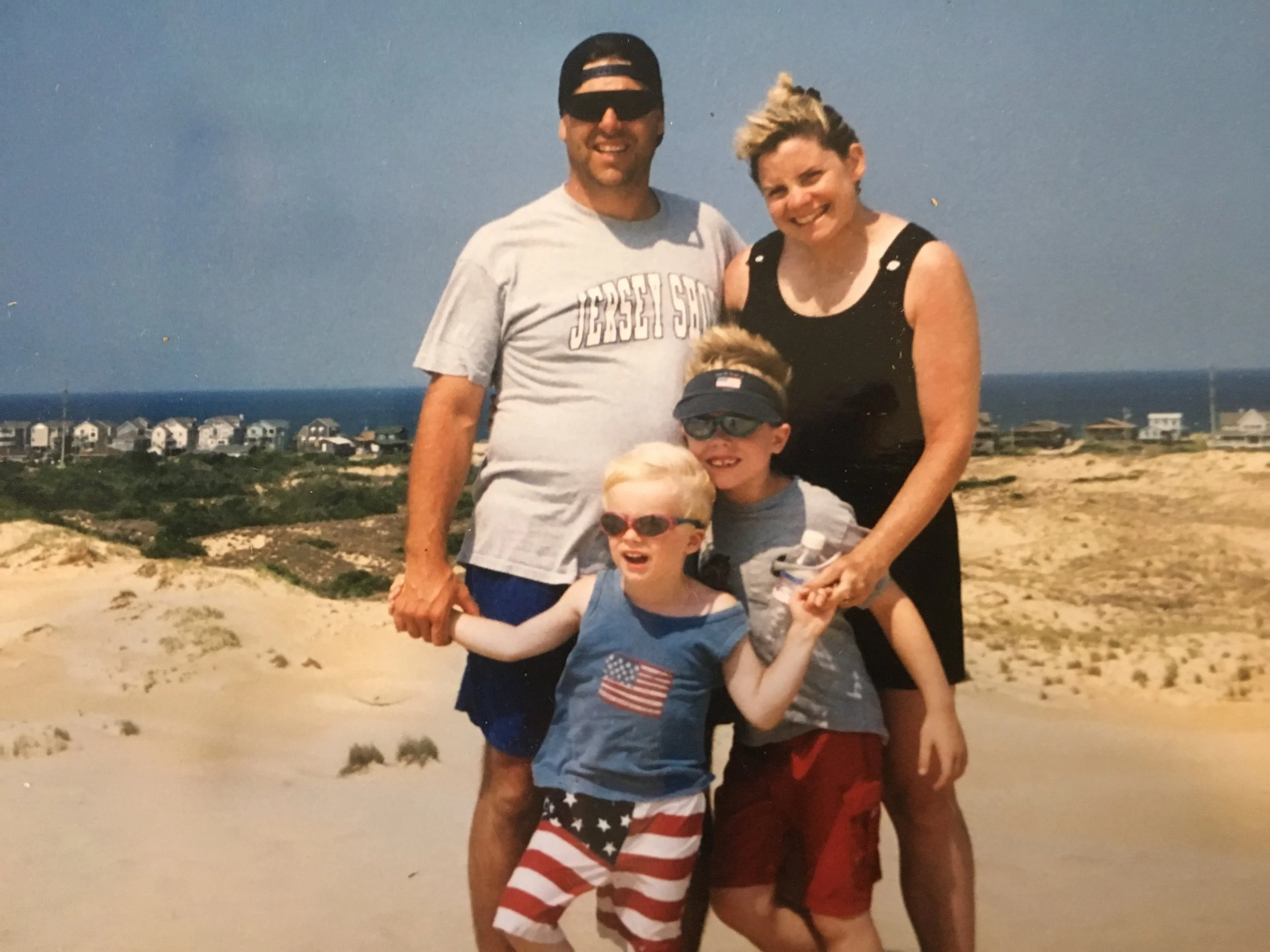 A family of four at a beach with sand dunes and houses in the background. The father is wearing a gray T-shirt and a black cap backward. The mother is dressed in a sleeveless black dress. The two children, both boys, are wearing sunglasses and casual summer clothes, with one in a blue tank top with an American flag and the other in a gray T-shirt. They are smiling and standing close together, enjoying a sunny day at the beach.