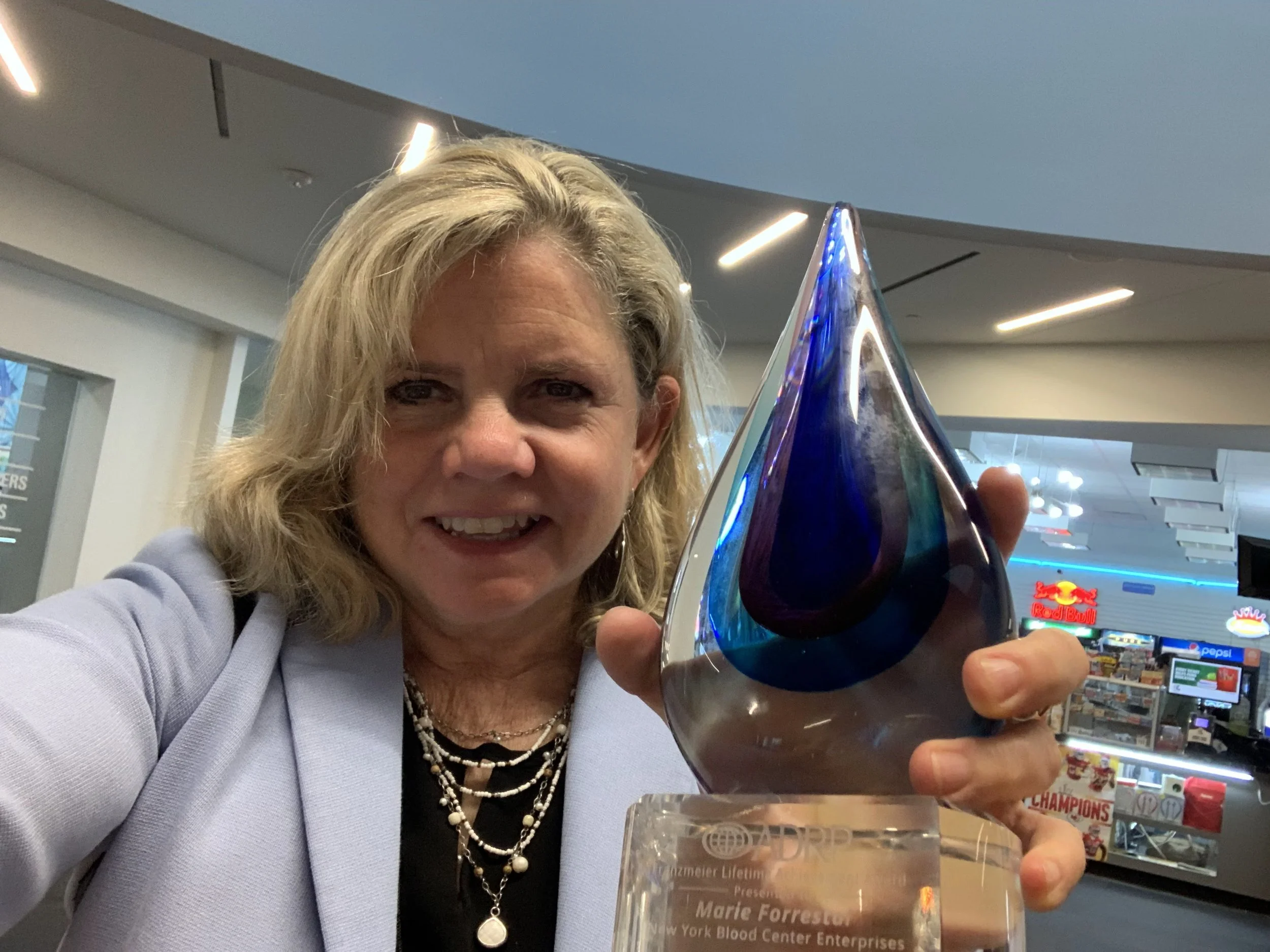A woman with blonde hair takes a selfie while holding an award trophy at an indoor location, with a background that includes a food concession stand and bright signage.