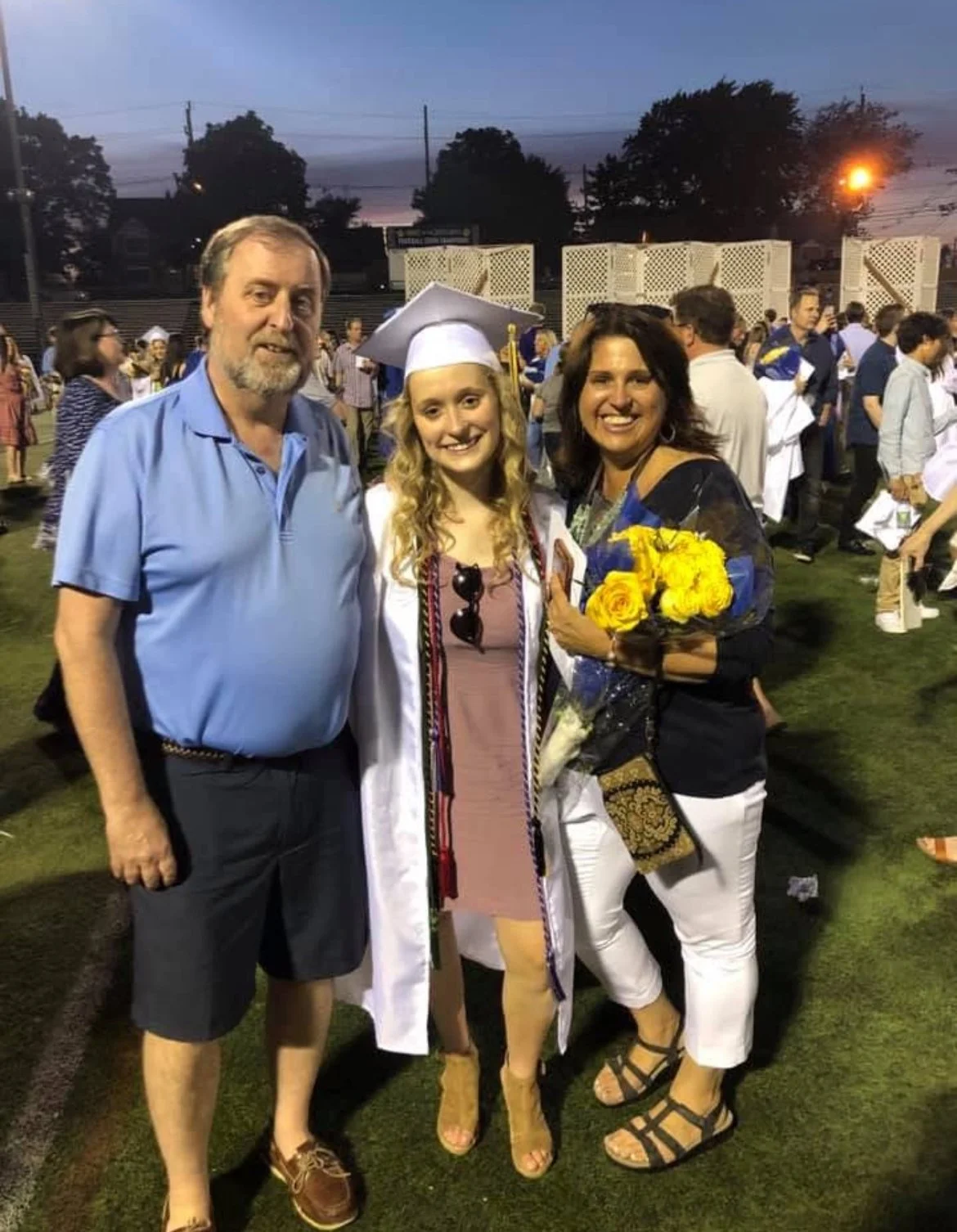 A young woman in a graduation gown and cap, smiling, standing between two adults, holding a bouquet of yellow flowers, at an outdoor graduation ceremony during sunset.