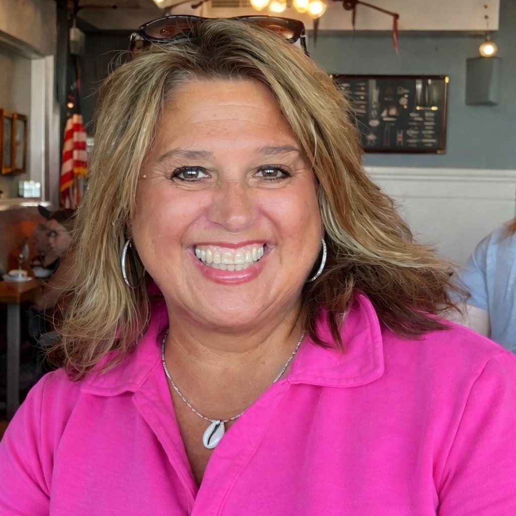 A woman in a pink shirt smiling at the camera in a restaurant or cafe with an American flag in the background.