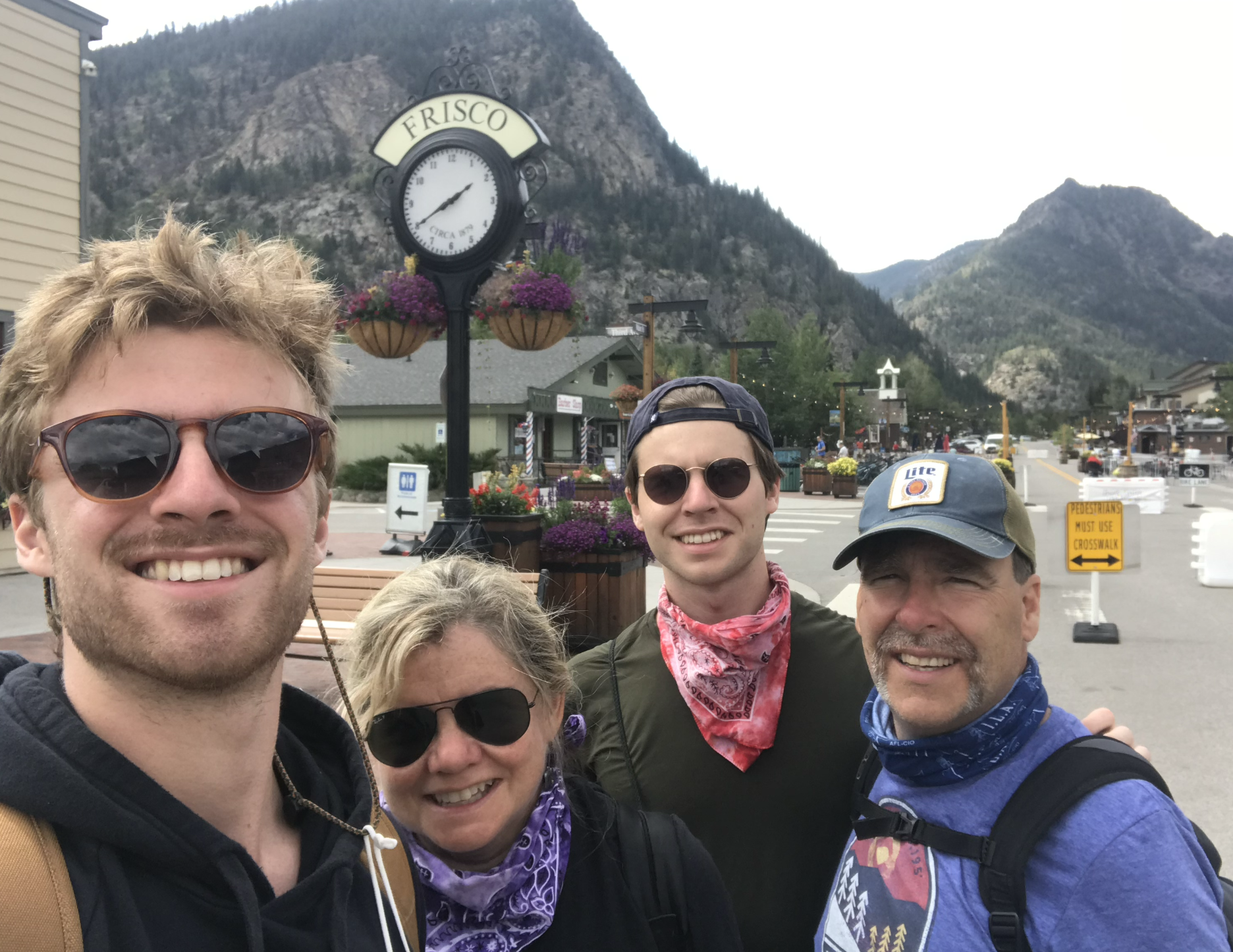 Four smiling friends are taking a selfie outdoors in a mountain town, with a sign reading 'FRISCO' and a clock behind them, surrounded by mountains and small shops.