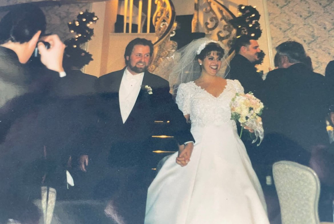 A bride and groom holding hands and smiling during their wedding reception, surrounded by guests.
