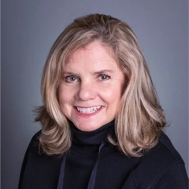 A woman with shoulder-length blonde hair and a big smile, wearing a black hoodie, against a gray background.