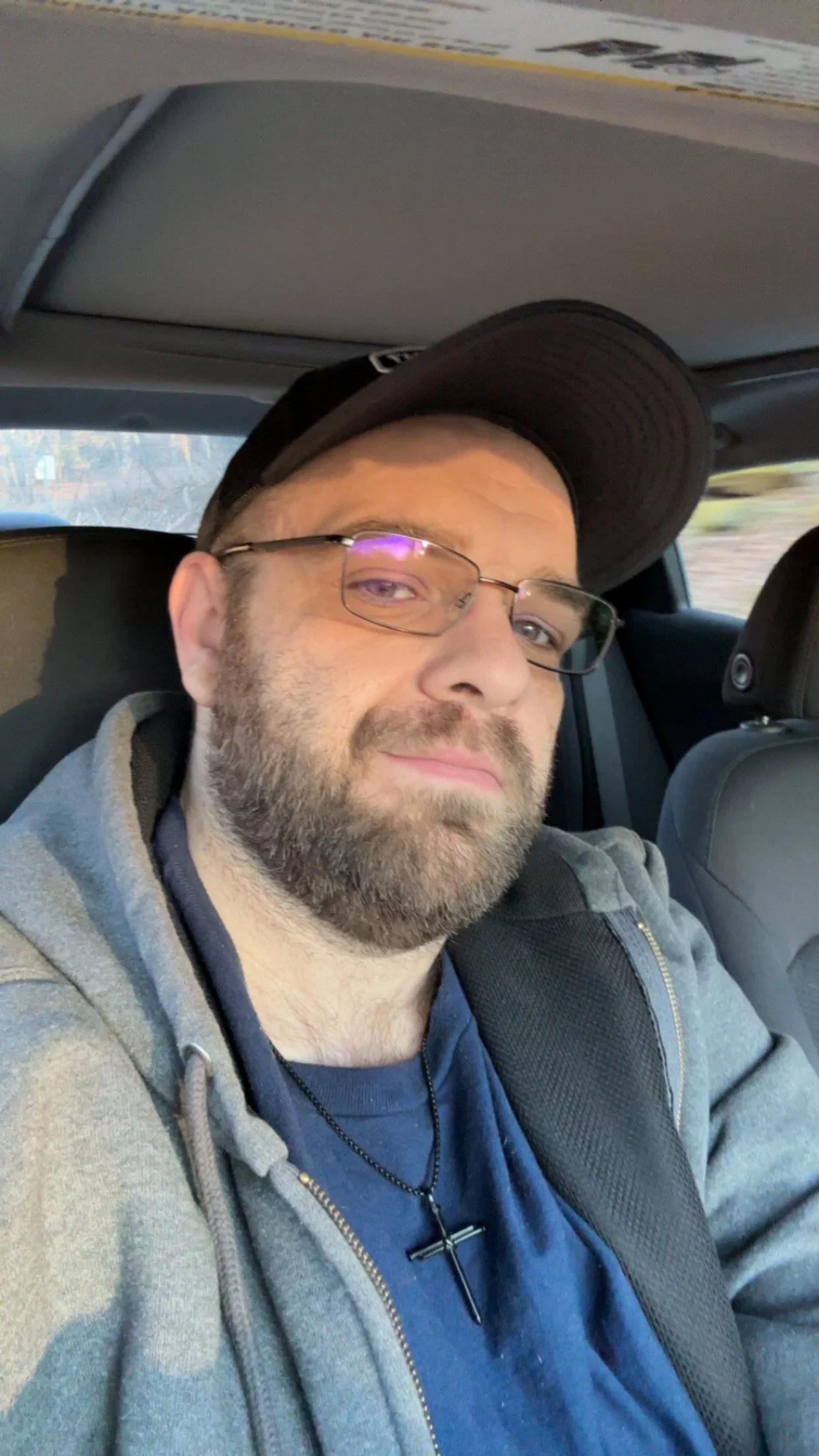 A man with glasses, a beard, and a baseball cap sitting in a car, smiling at the camera.