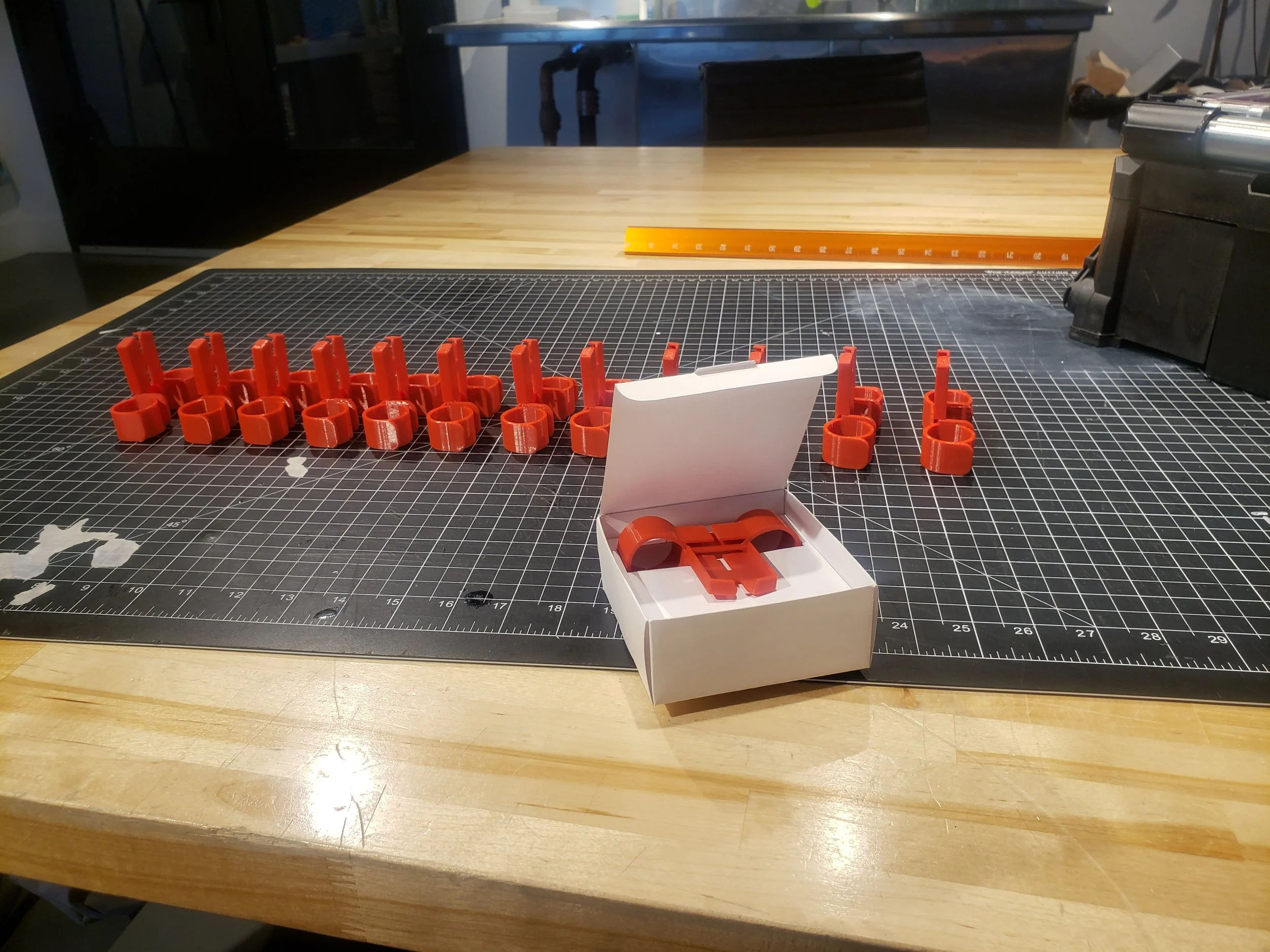 A black cutting mat on a wooden table with small orange plastic parts arranged in two rows, a white box containing additional orange parts, and a metal ruler in the background.