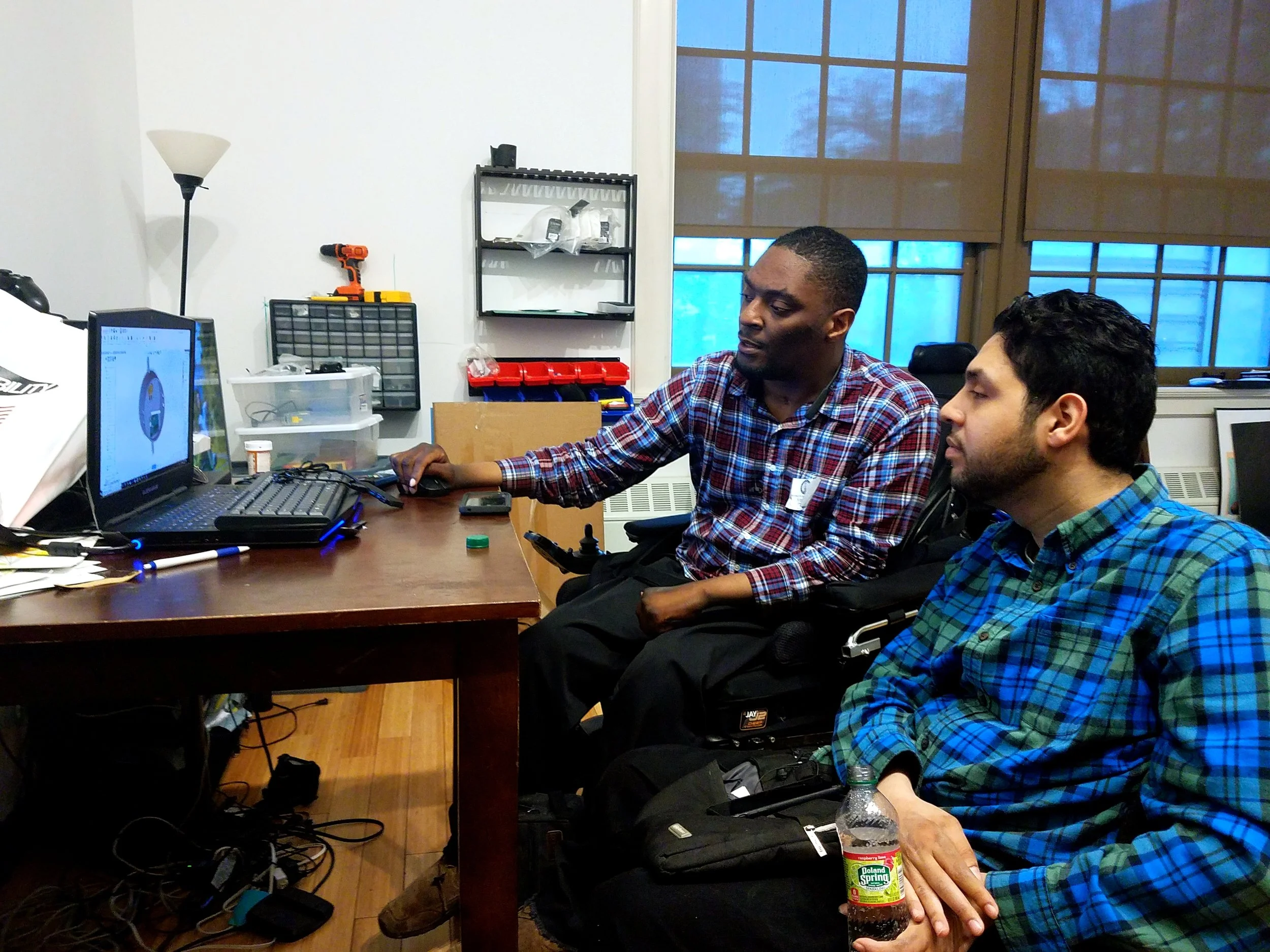 Two men sit at a desk, looking at a computer screen. One is in a wheelchair, and the other is holding a bottle of soda, both focused on the monitor.