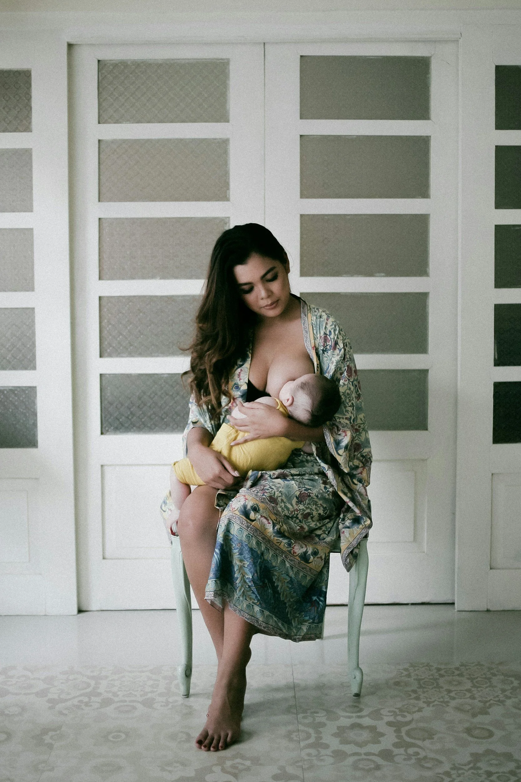 A woman with long dark hair sitting on a white chair, breastfeeding a baby in a yellow outfit, indoors in front of a white-paneled wall with textured glass windows.