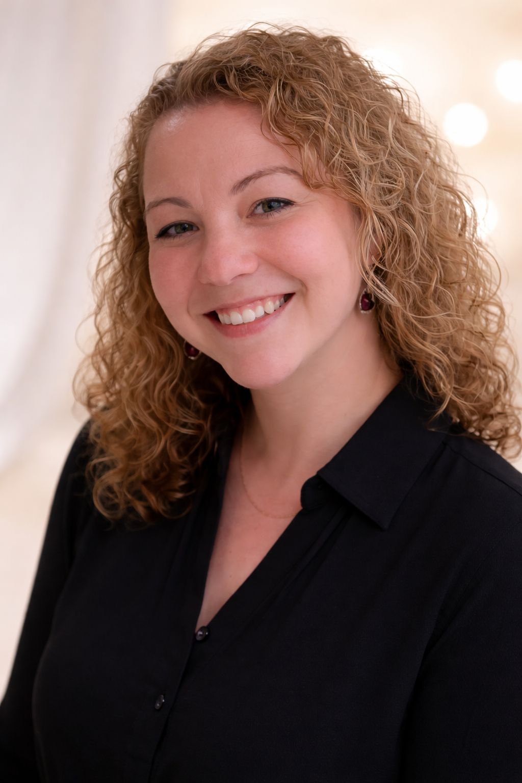 A smiling woman with curly blonde hair, wearing a black shirt and earrings, standing indoors.